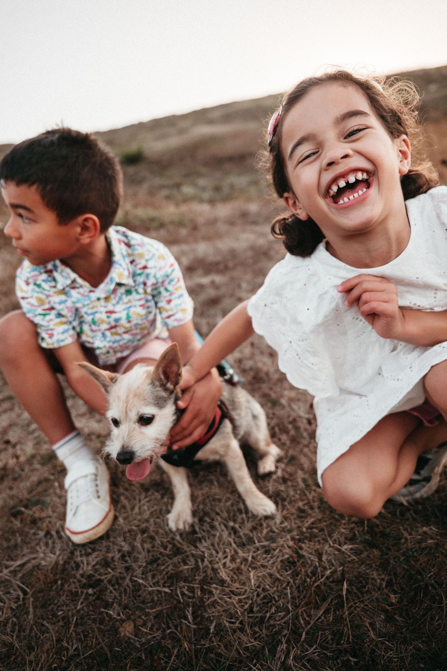 Two children laugh and pet a small dog outdoors on dry grass at sunset.