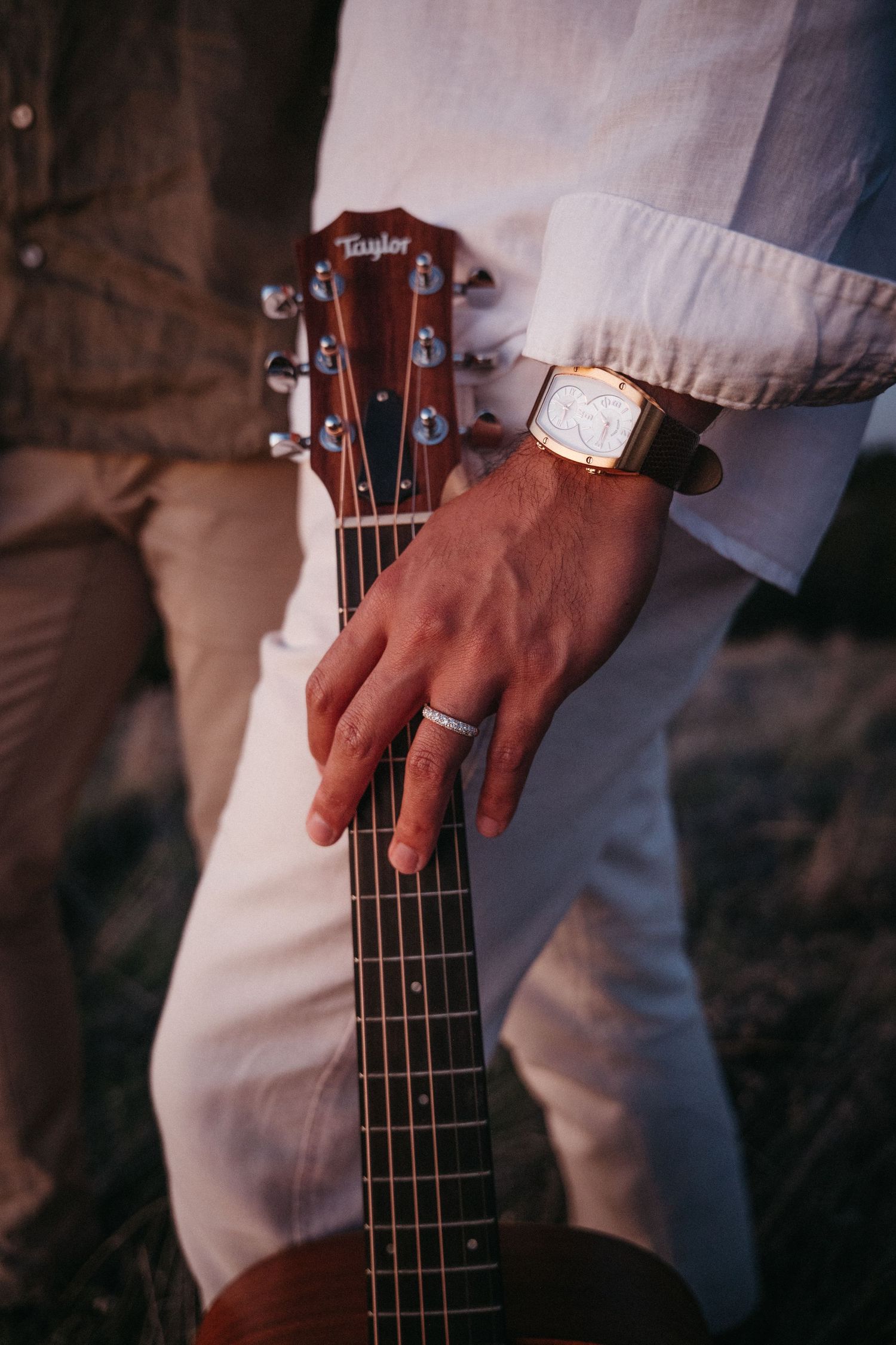 Close-up of a hand with a ring and gold watch resting on a Taylor acoustic guitar neck outdoors.