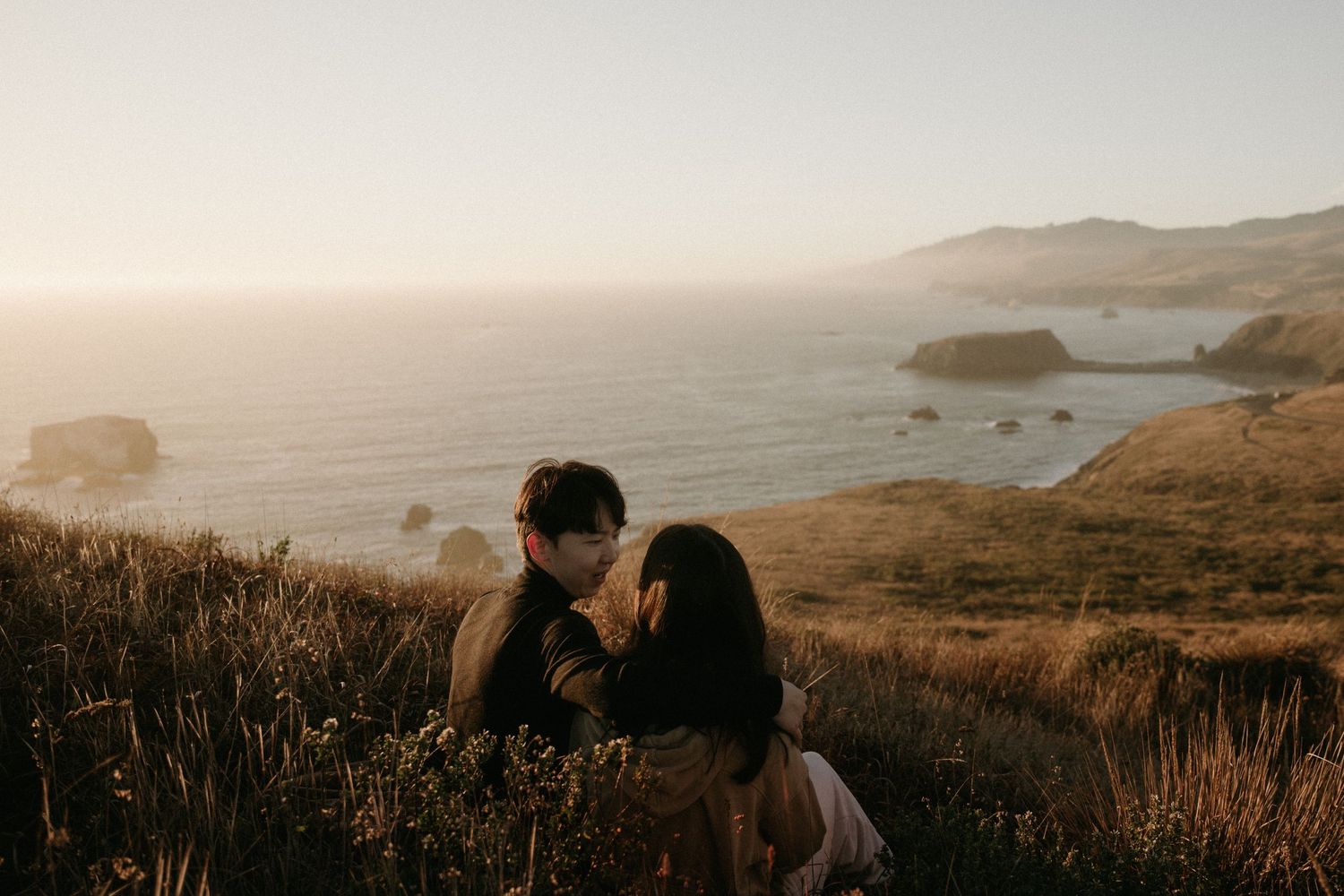 Couple sits close together on a grassy coastal hillside at golden hour, overlooking the ocean and rocky sea stacks.