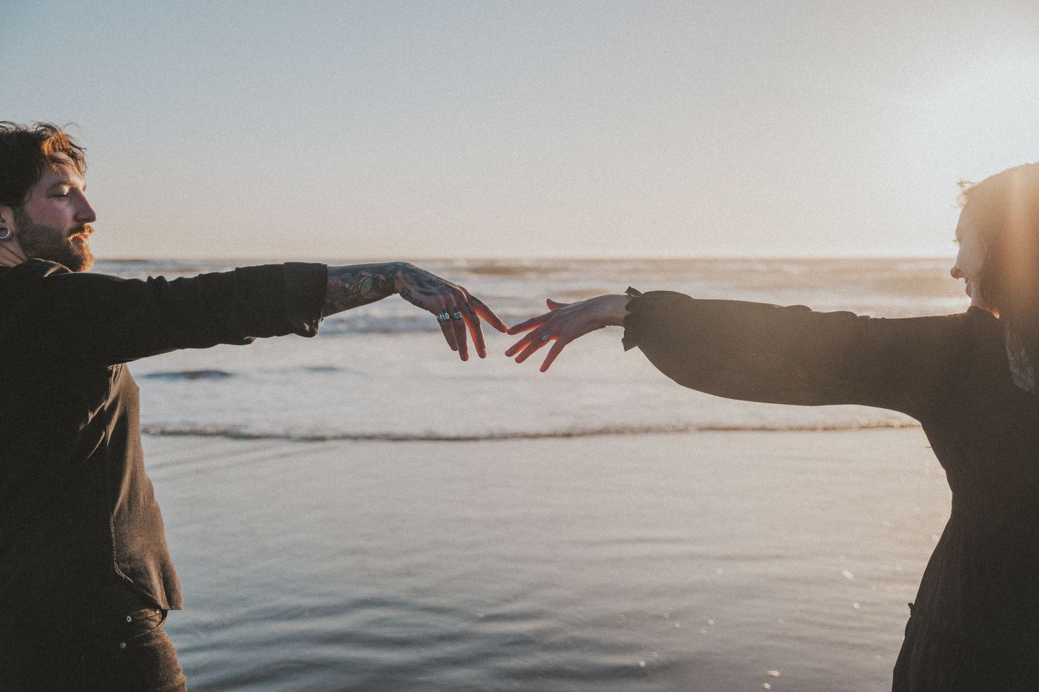 Two people reaching toward each other on a sunlit beach with ocean waves in the background.