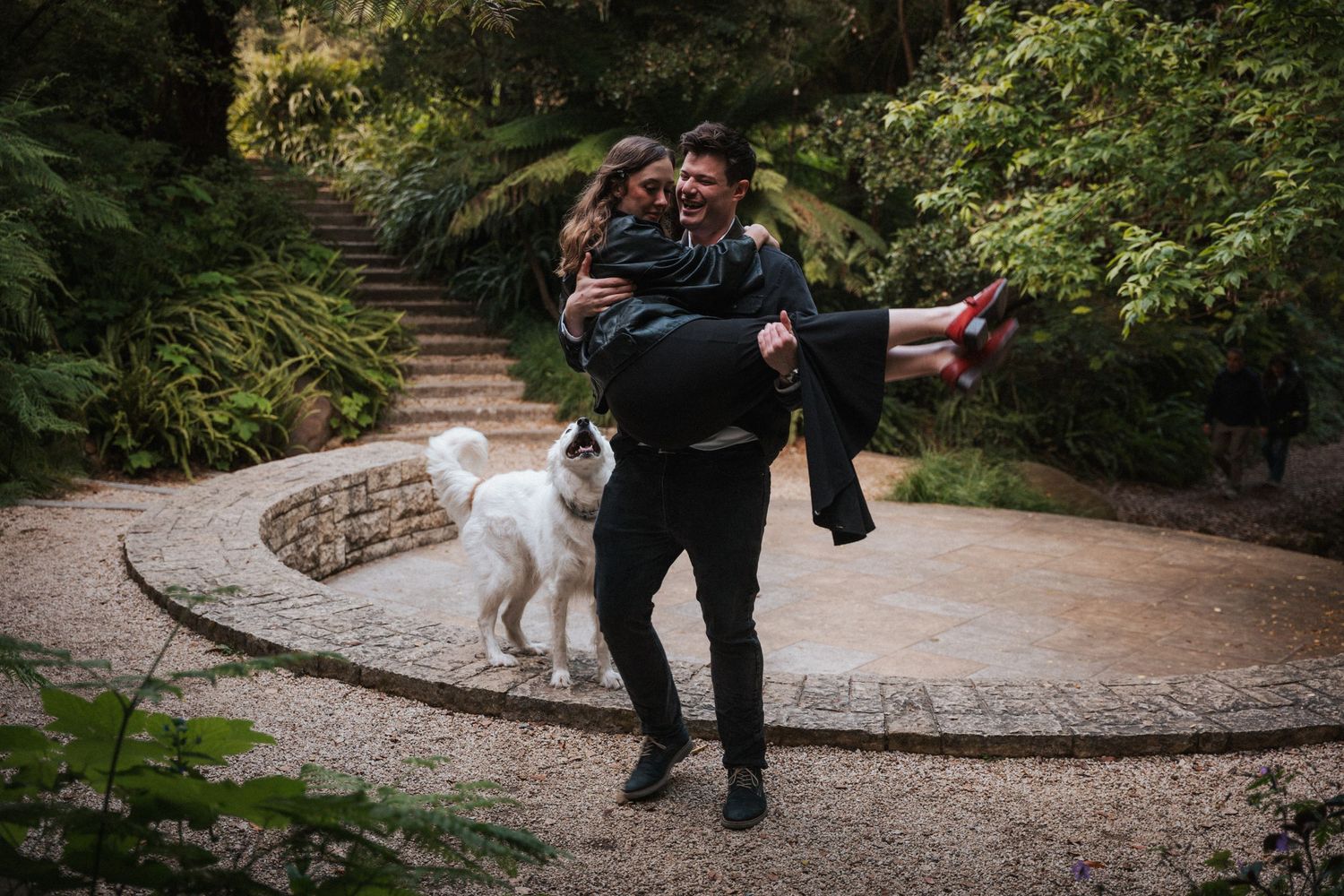 A man carries a woman in his arms while a white dog walks beside them in a lush garden setting.