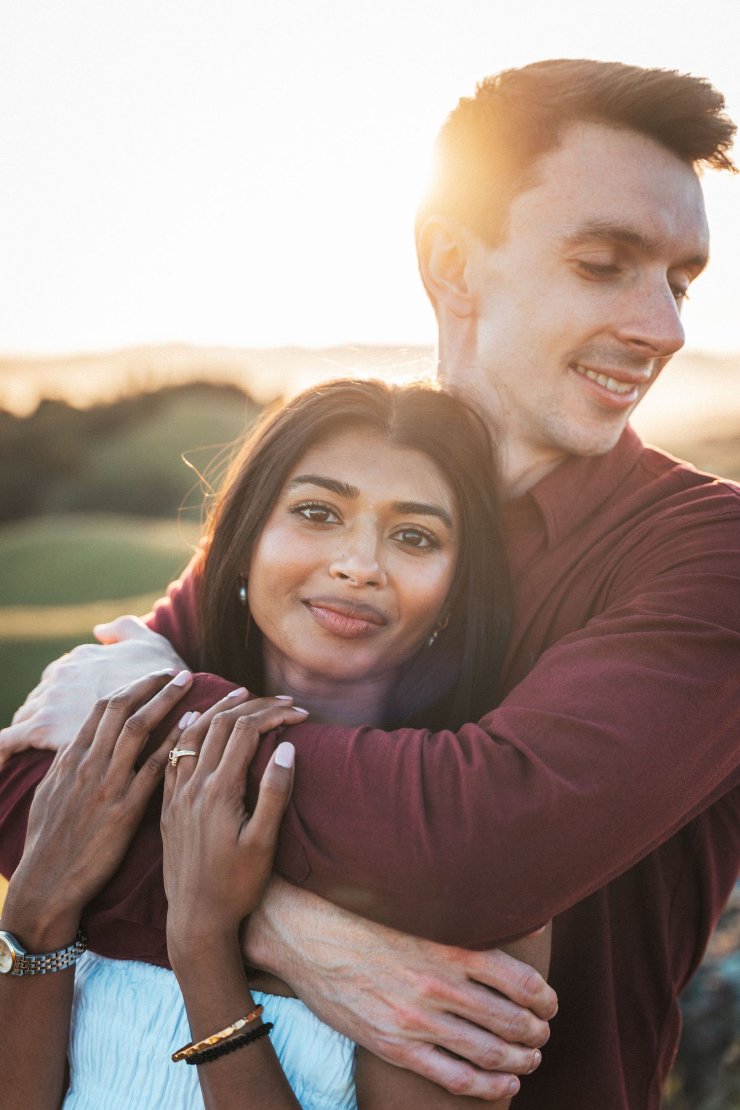 Happy interracial couple embracing at sunset in scenic outdoor setting, woman showing engagement ring.