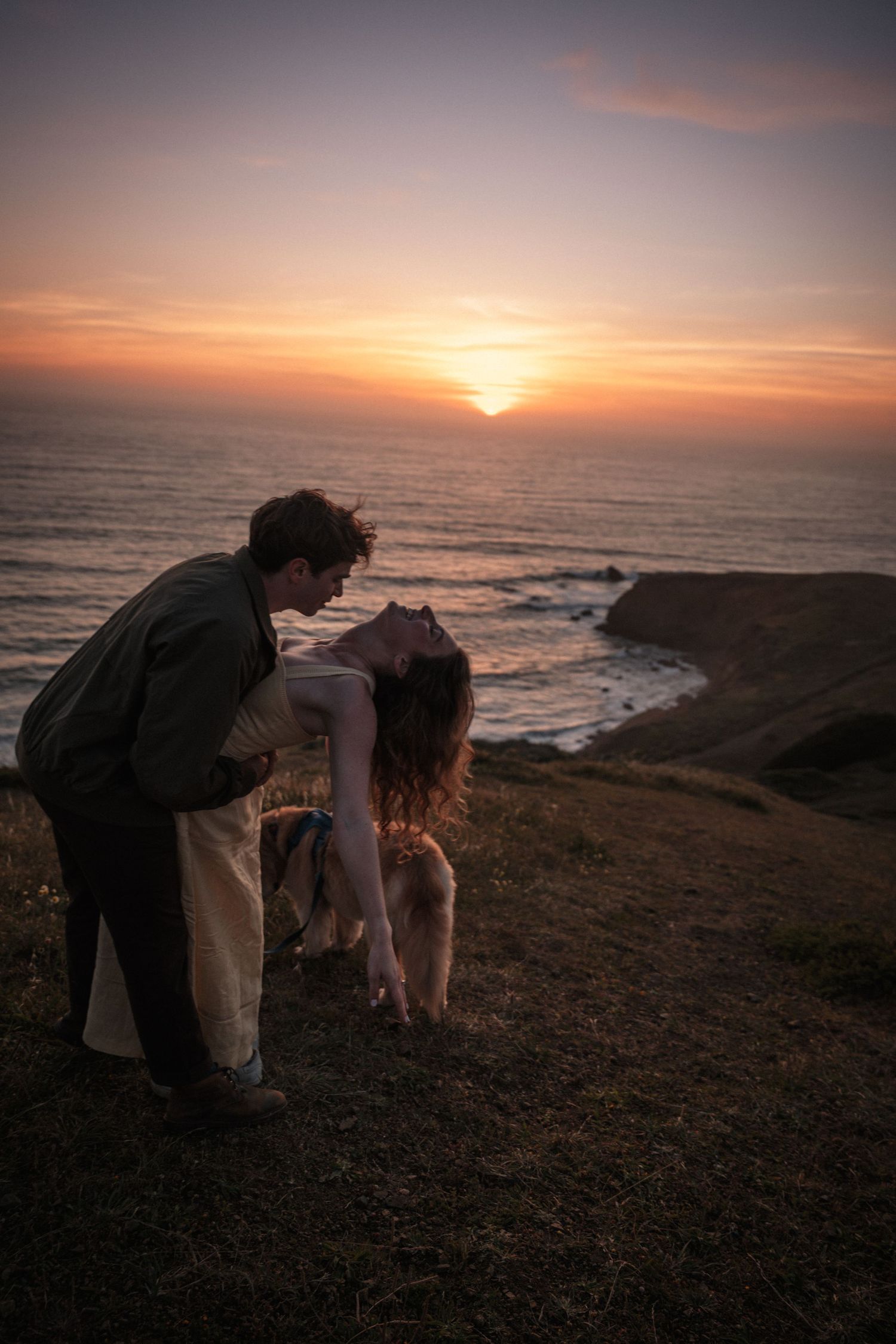 Romantic couple sharing a tender moment on a coastal cliff at sunset with ocean views.