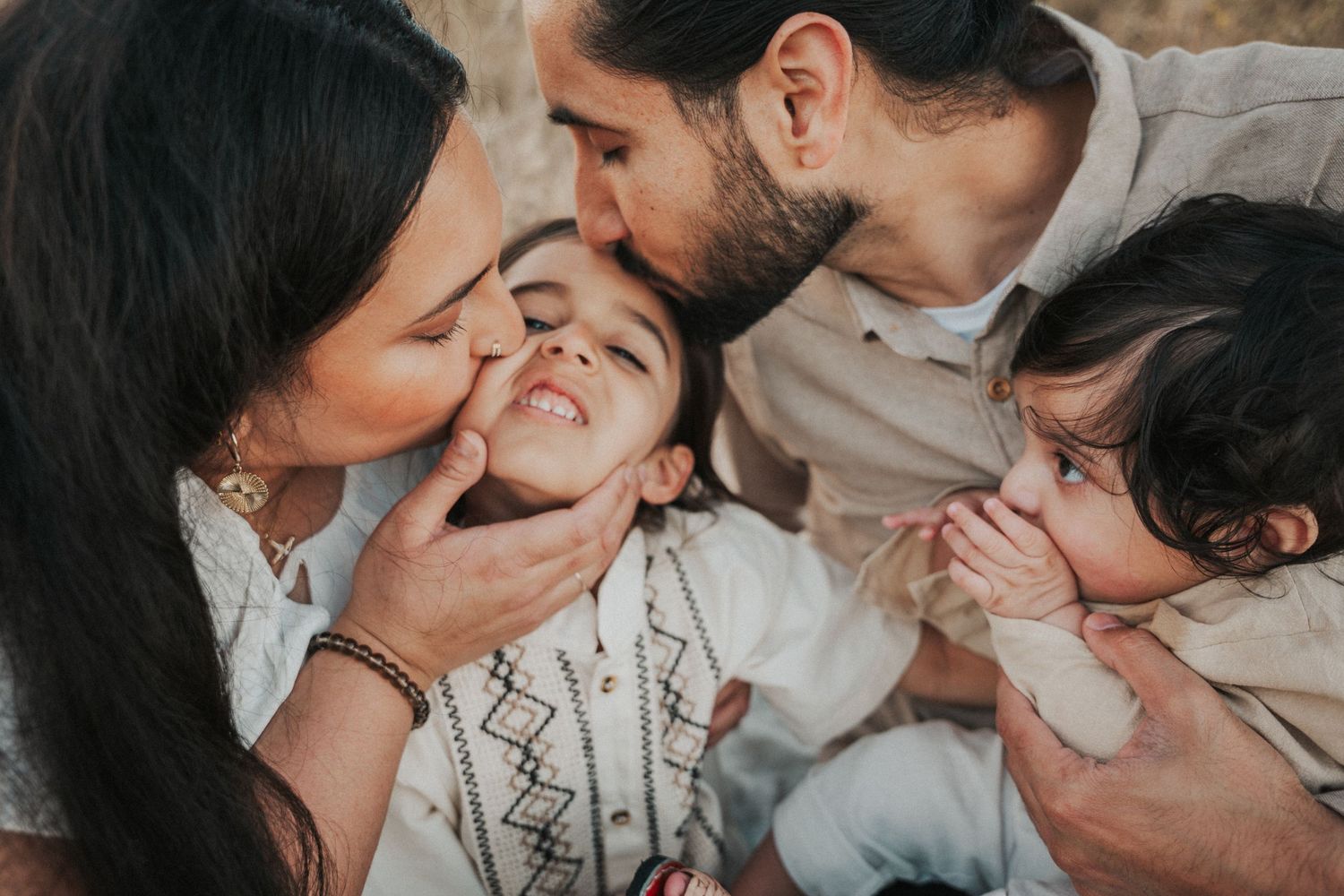 Happy family with parents kissing young children on cheeks outdoors in a warm, candid portrait.
