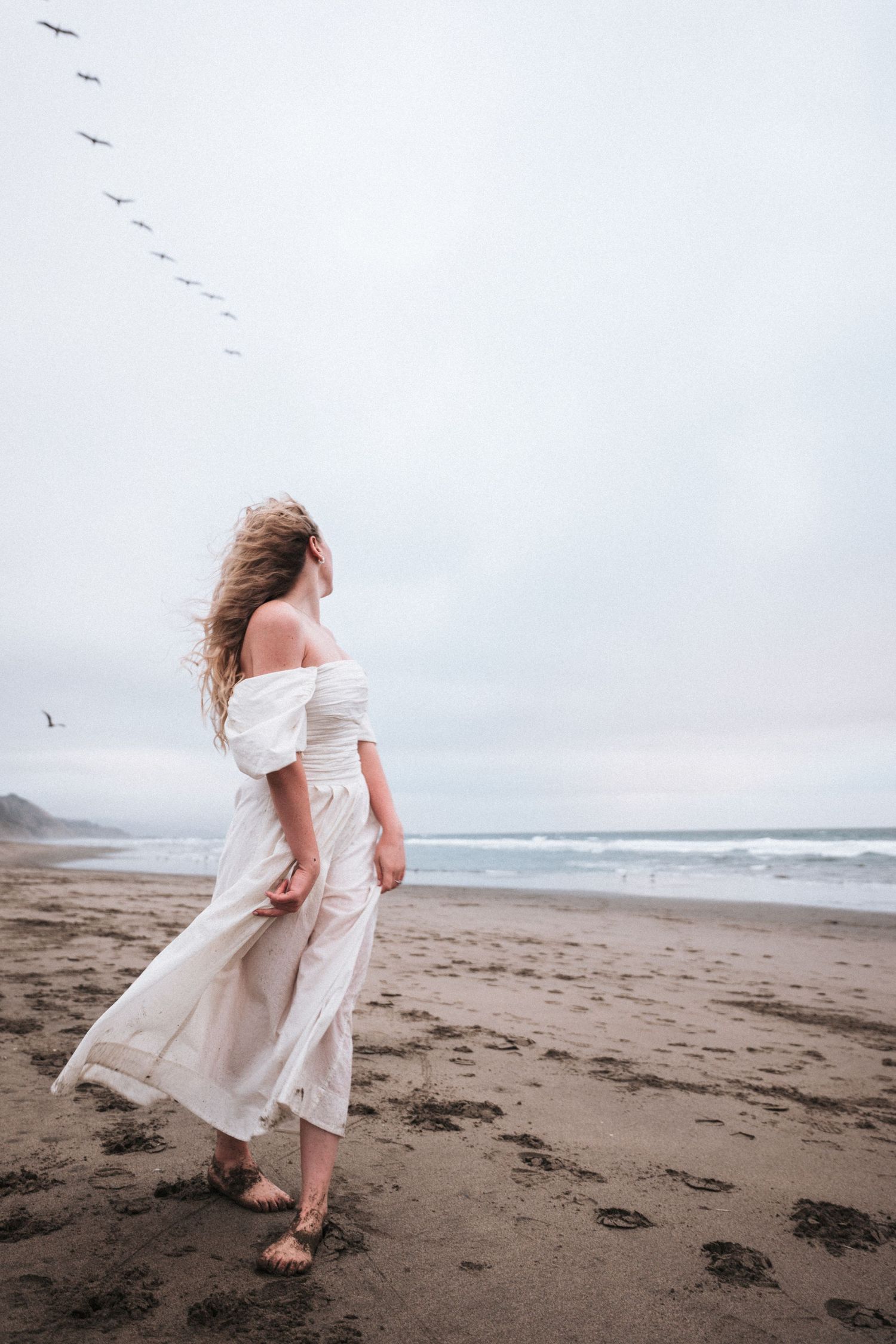 Woman in flowing white dress walking barefoot on sandy beach with birds flying overhead.