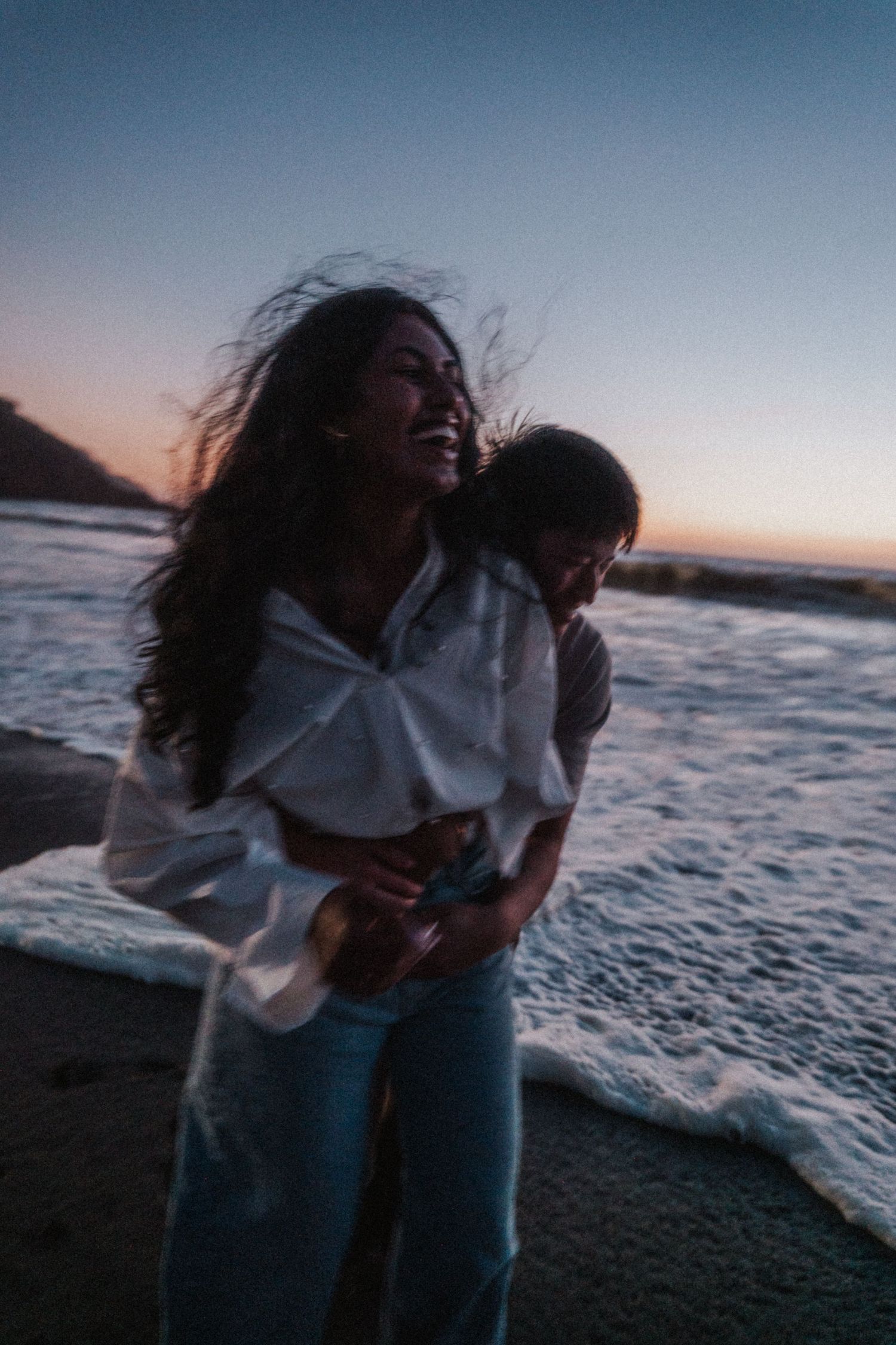 Joyful woman in white shirt and jeans laughing as man hugs her from behind on beach at sunset.