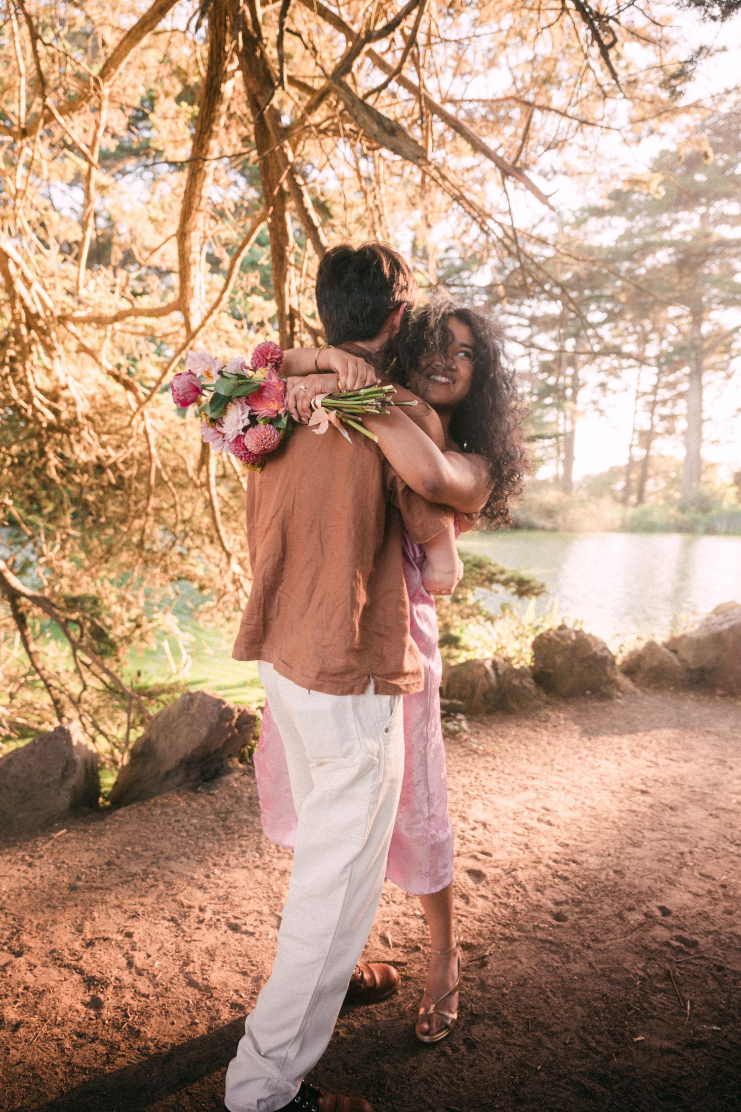 Couple embracing outdoors near a lake, woman smiling holding pink flowers, surrounded by autumn trees in warm sunlight.