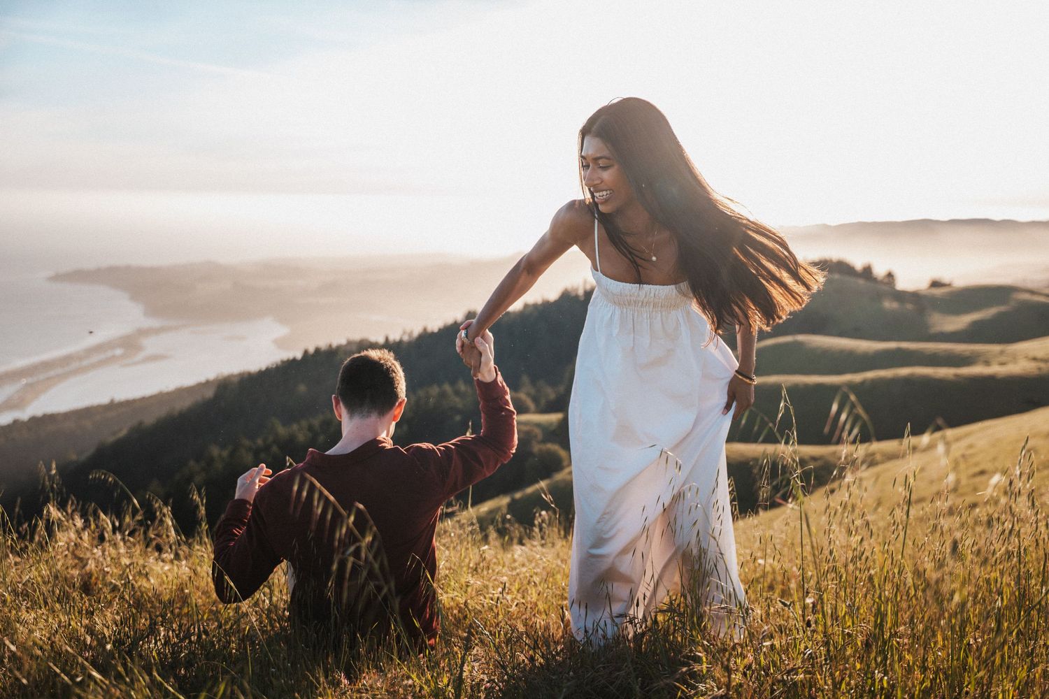 Couple dancing on a sunlit hillside overlooking the ocean, woman in white dress spinning gracefully at golden hour.