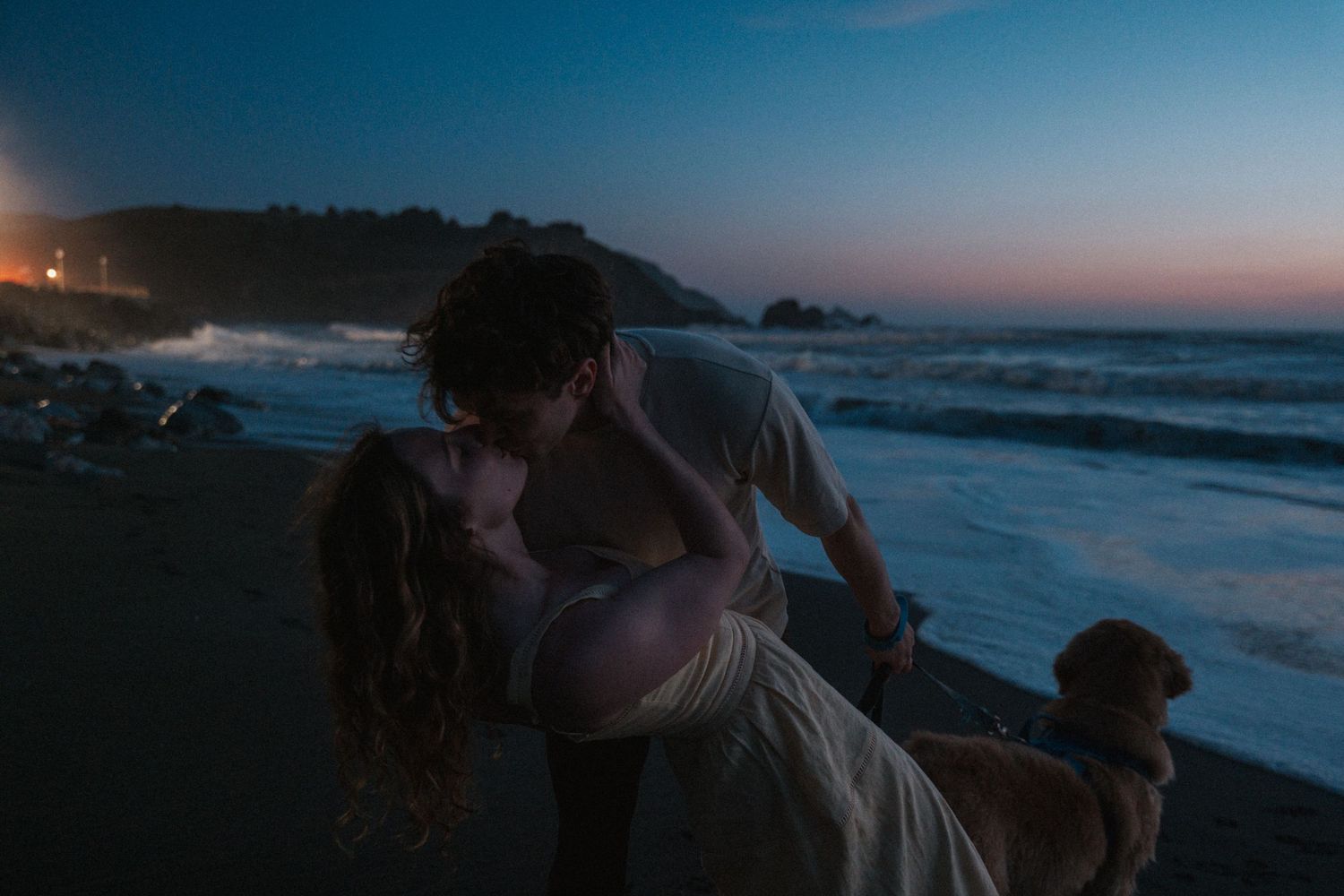 Couple sharing a romantic kiss on a dark beach at night with their dog nearby.