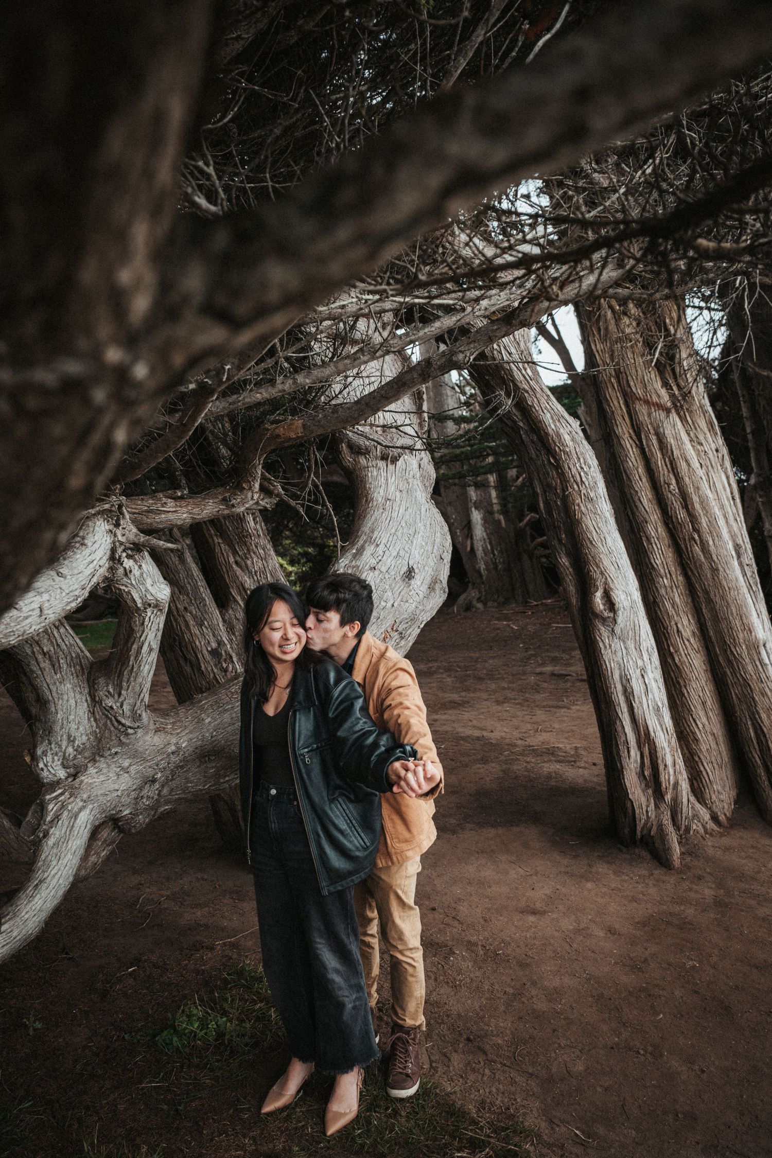 Couple embracing and kissing under twisted cypress trees in a moody forest setting.