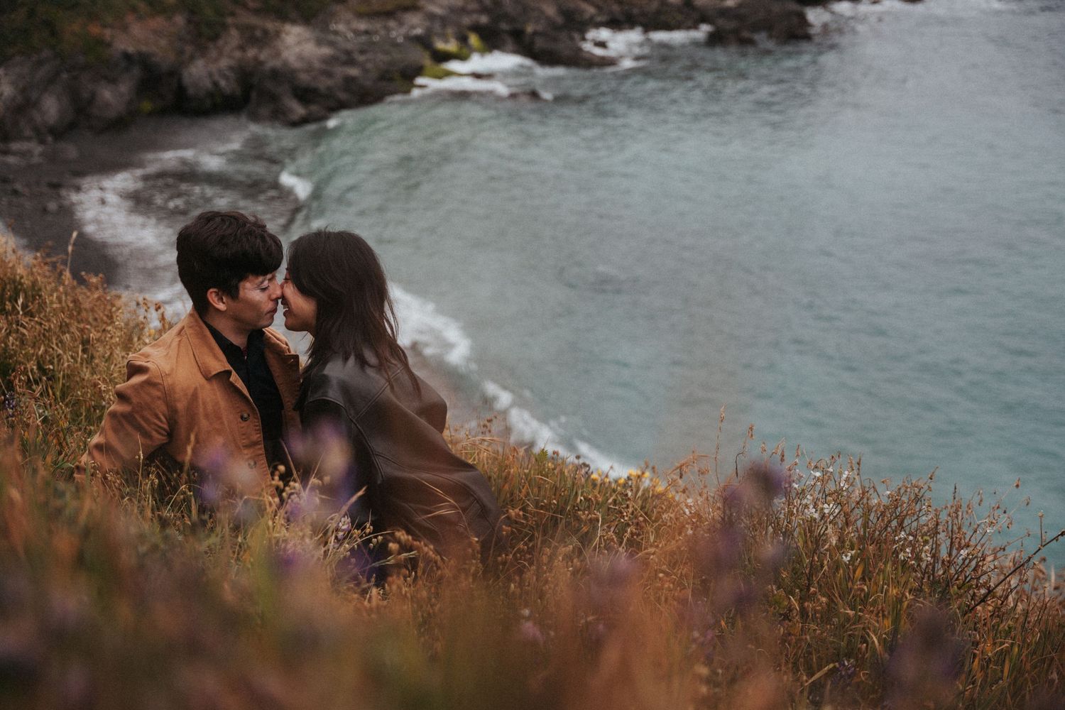 Couple sharing a romantic moment on a coastal cliff overlooking turquoise ocean waters surrounded by wildflowers.