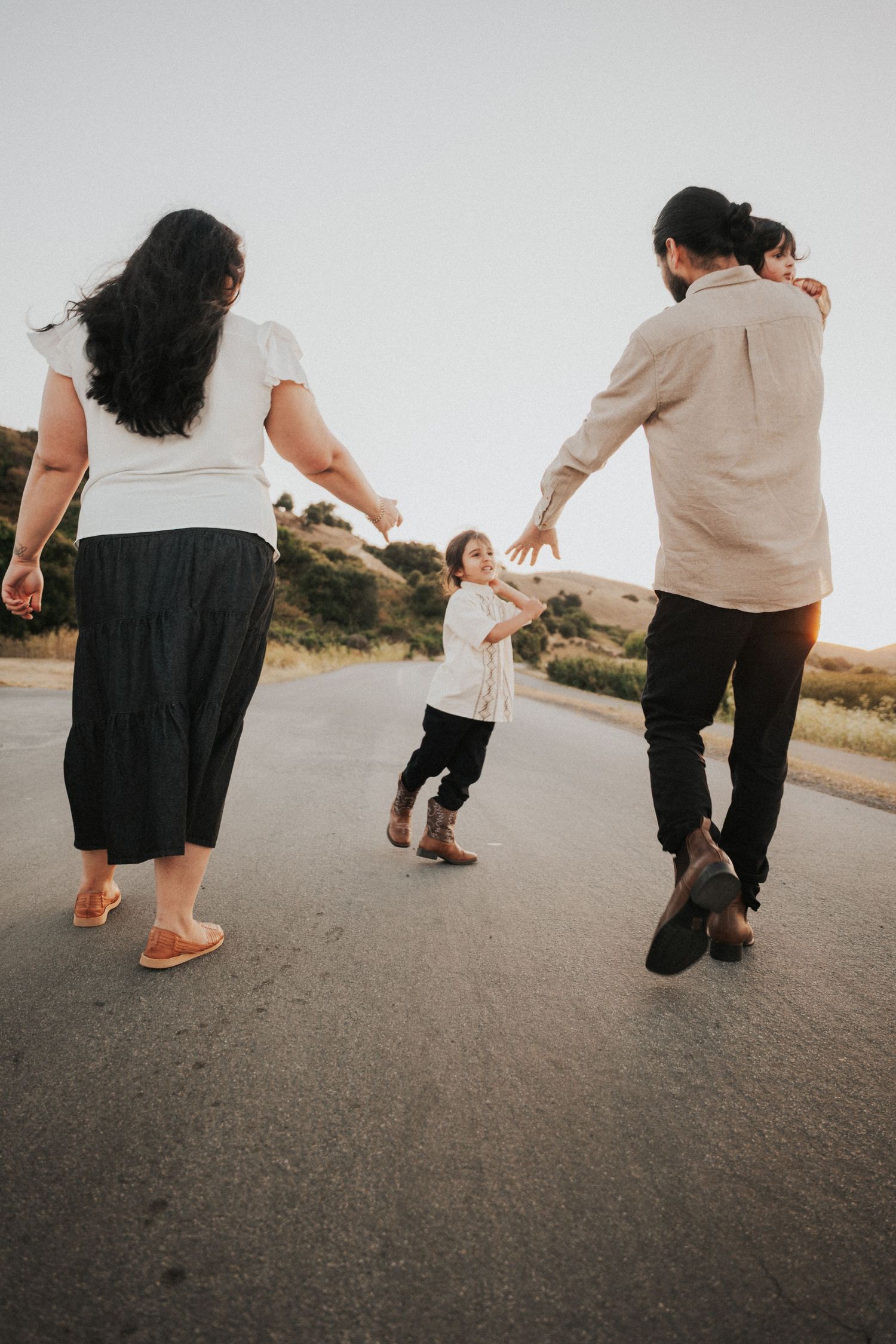 Family walking on rural road at sunset, parents reaching toward toddler standing between them.