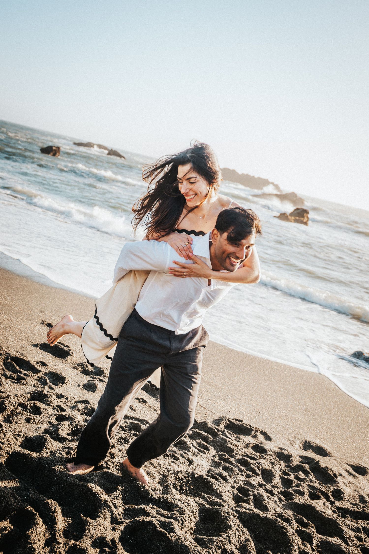 A joyful couple plays on a sandy beach, man carrying woman in his arms with ocean waves in the background.