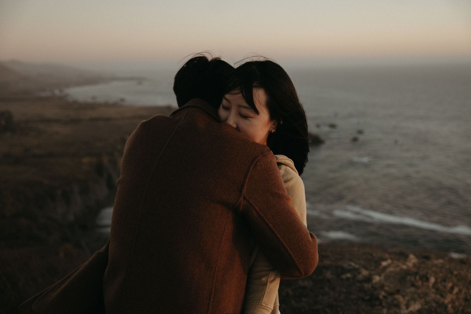 Couple embracing tightly on a coastal cliff at sunset, with the ocean and rugged shoreline stretching out behind them.
