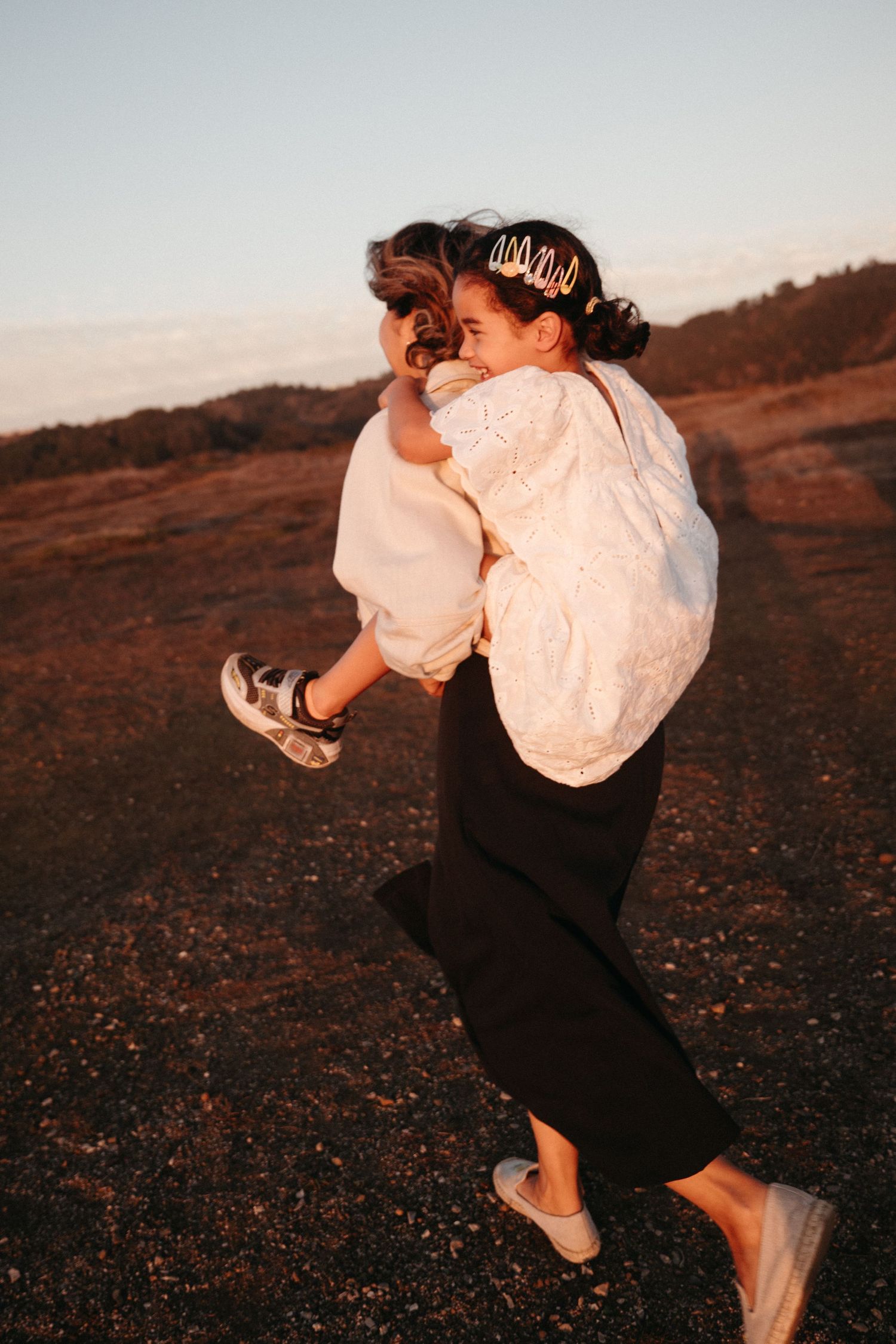 A woman lifts and hugs a small child in a white dress on a hilltop at dusk.