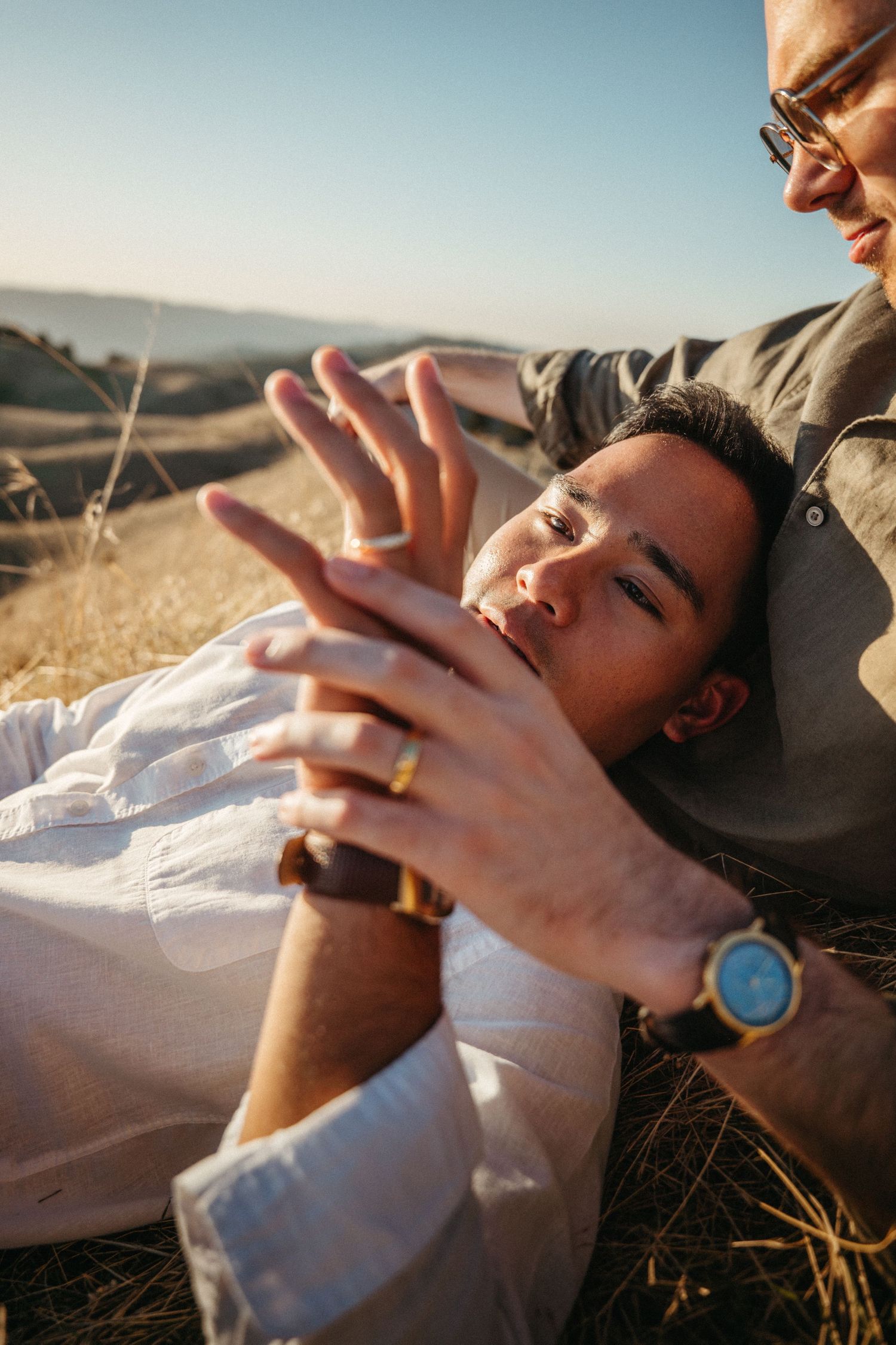 Two men relaxing outdoors on dry grass, one lying down admiring a gold ring on his hand at sunset.