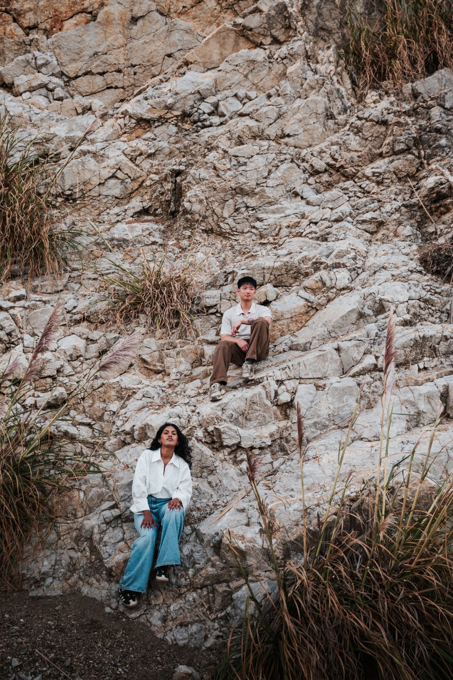 Couple posing on rocky cliff face with dry grass, woman in white shirt and blue pants sits below man on rocks.