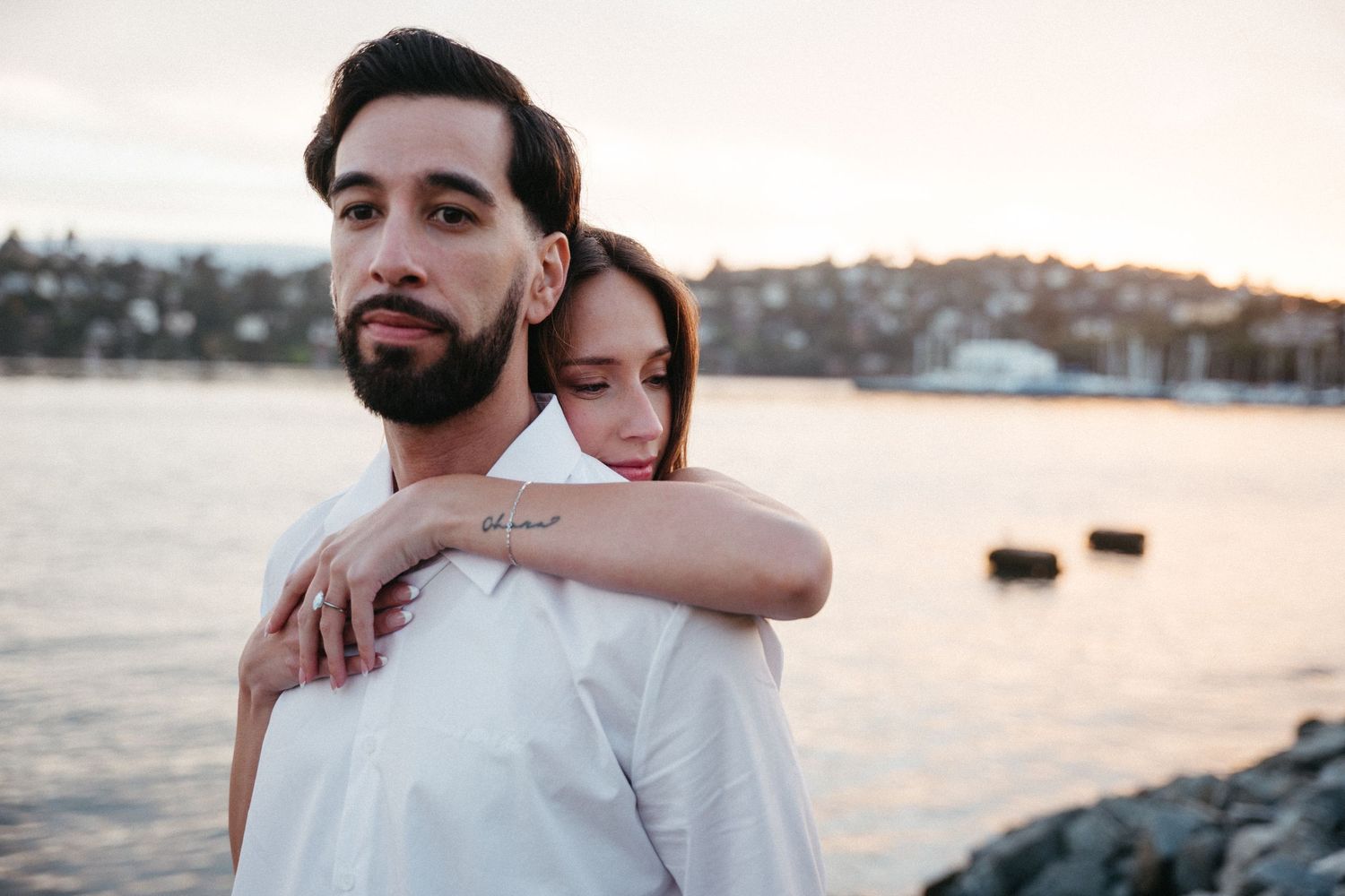 A couple poses near a waterfront at sunset, woman embracing man from behind, both wearing white.