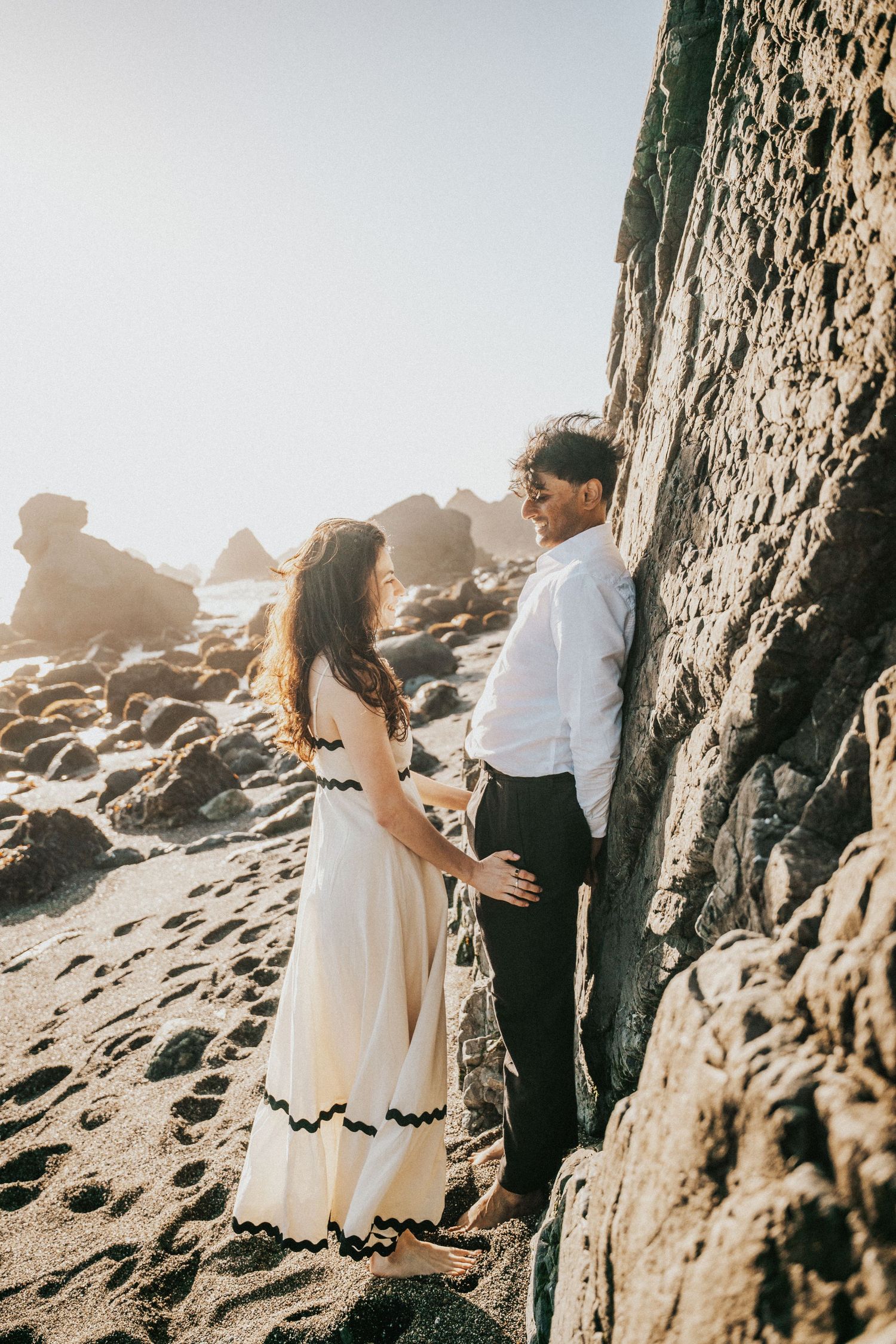 Couple holding hands near rocky cliffs on a sandy beach, woman in white dress, man in white shirt and dark pants.