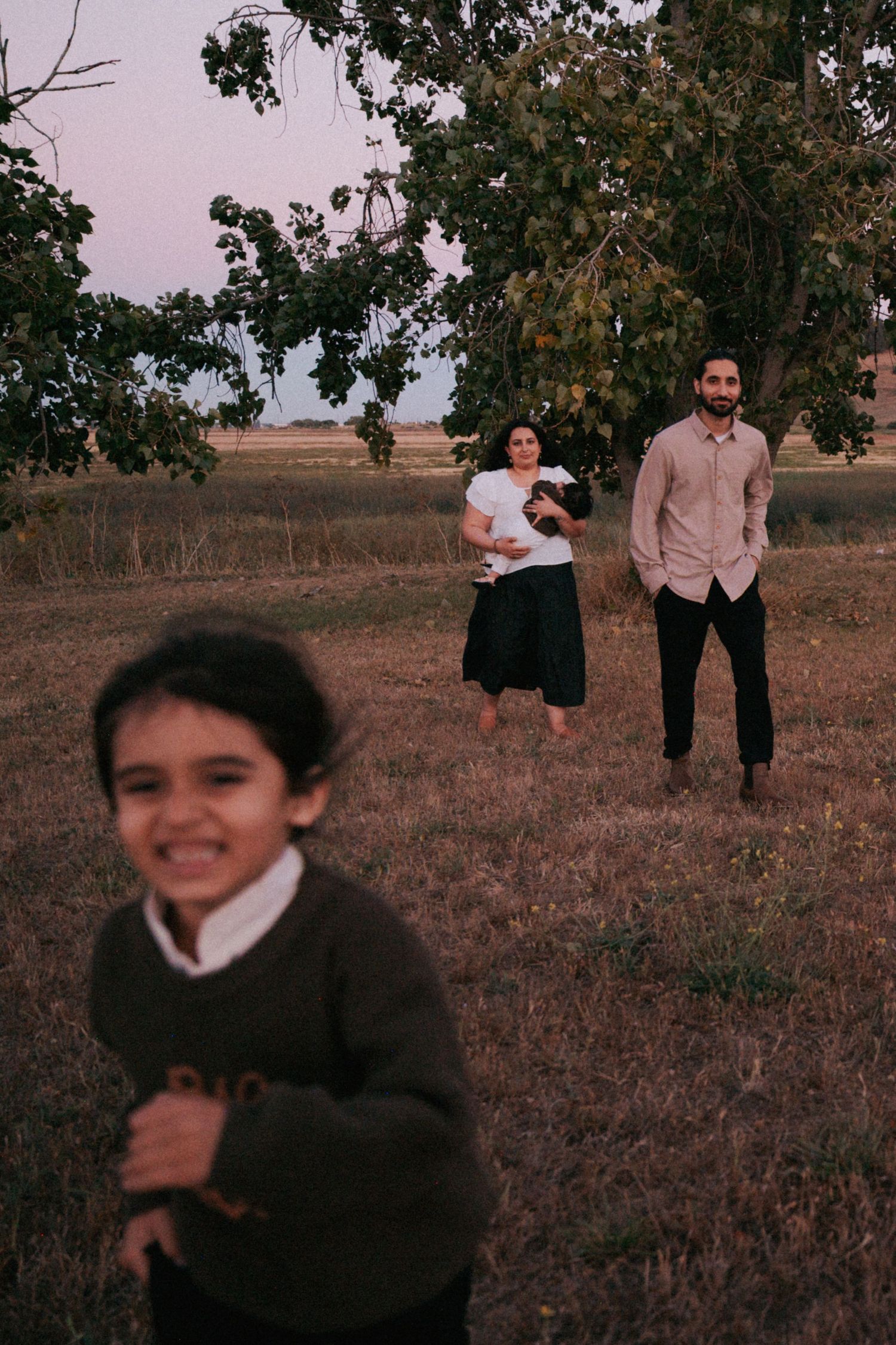 A young child runs toward the camera while parents stand with a baby near a tree in an open field at dusk.