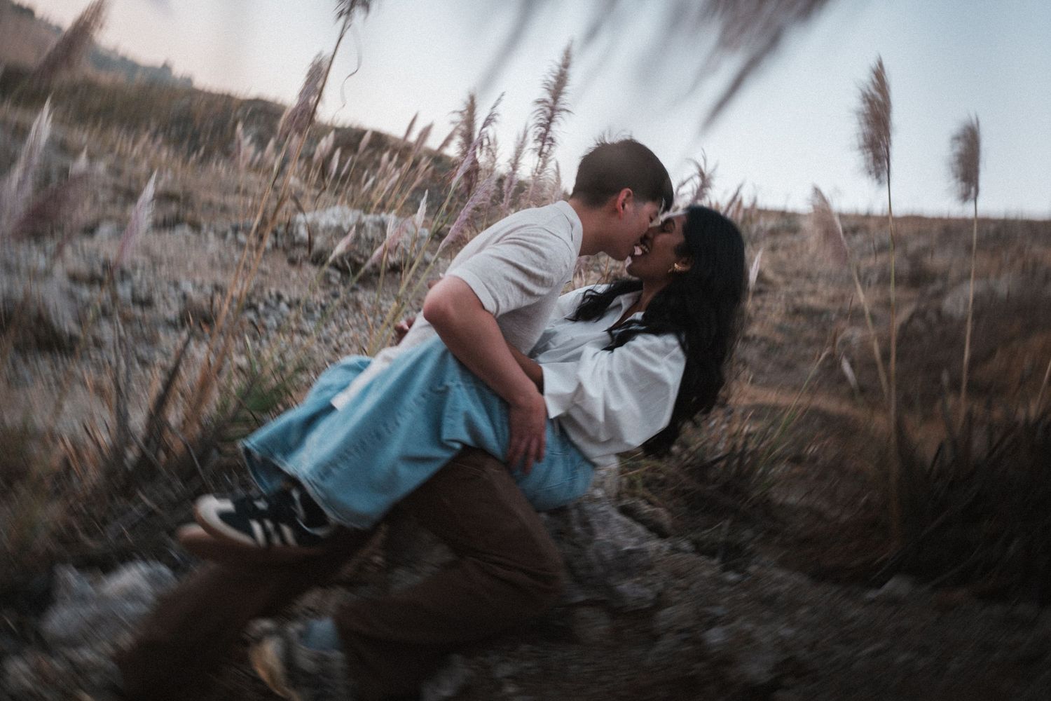 A couple sharing a romantic dip kiss in a golden field at dusk, surrounded by tall wild grasses.