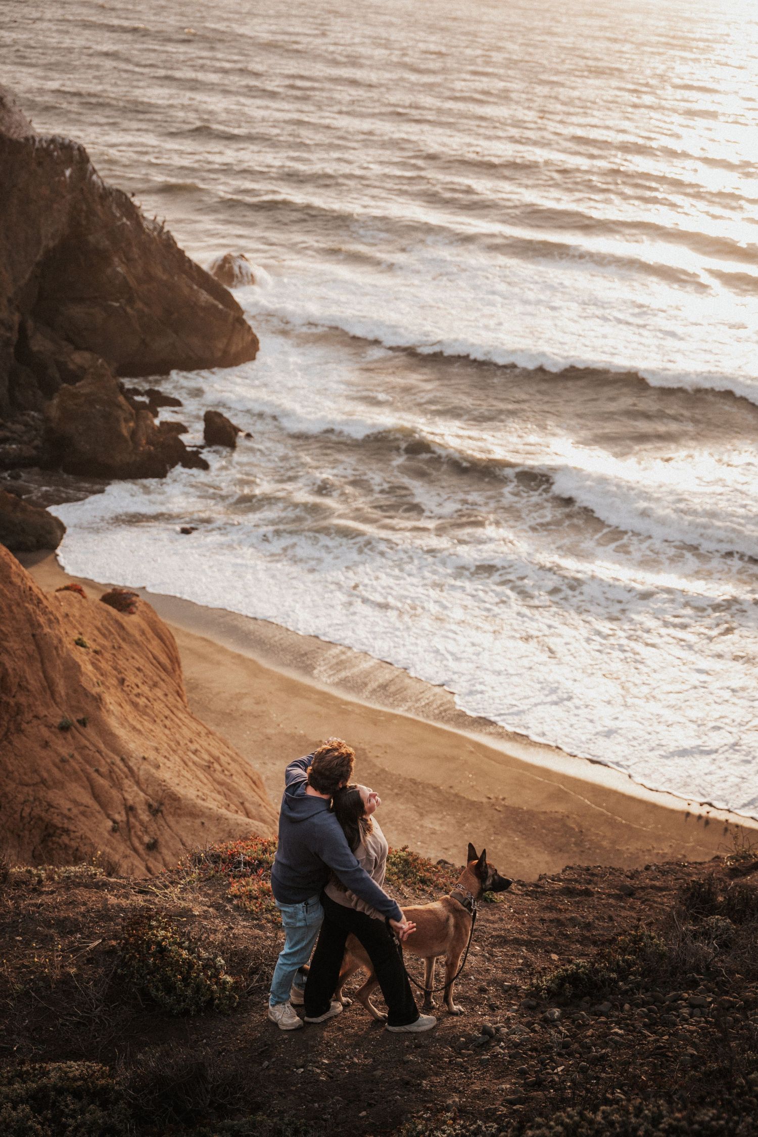 Couple embracing with a dog on a coastal cliff overlooking sandy beach and ocean waves at golden hour.