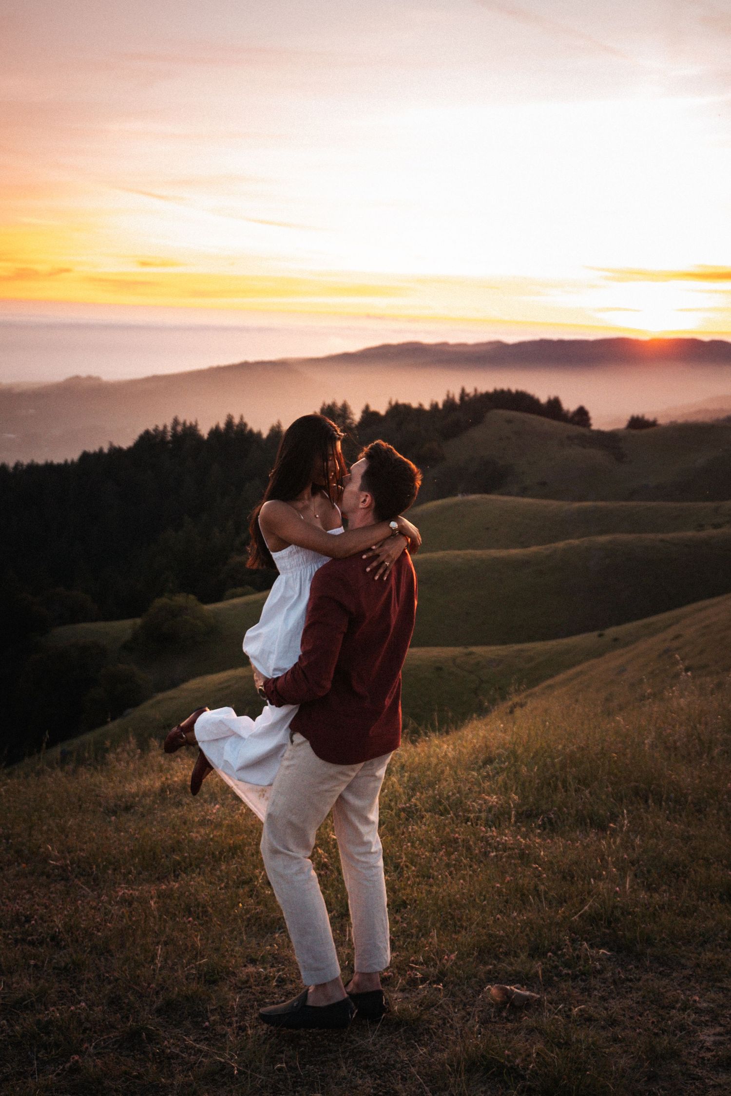 Couple sharing romantic kiss on green hillside with stunning layered mountain sunset backdrop.