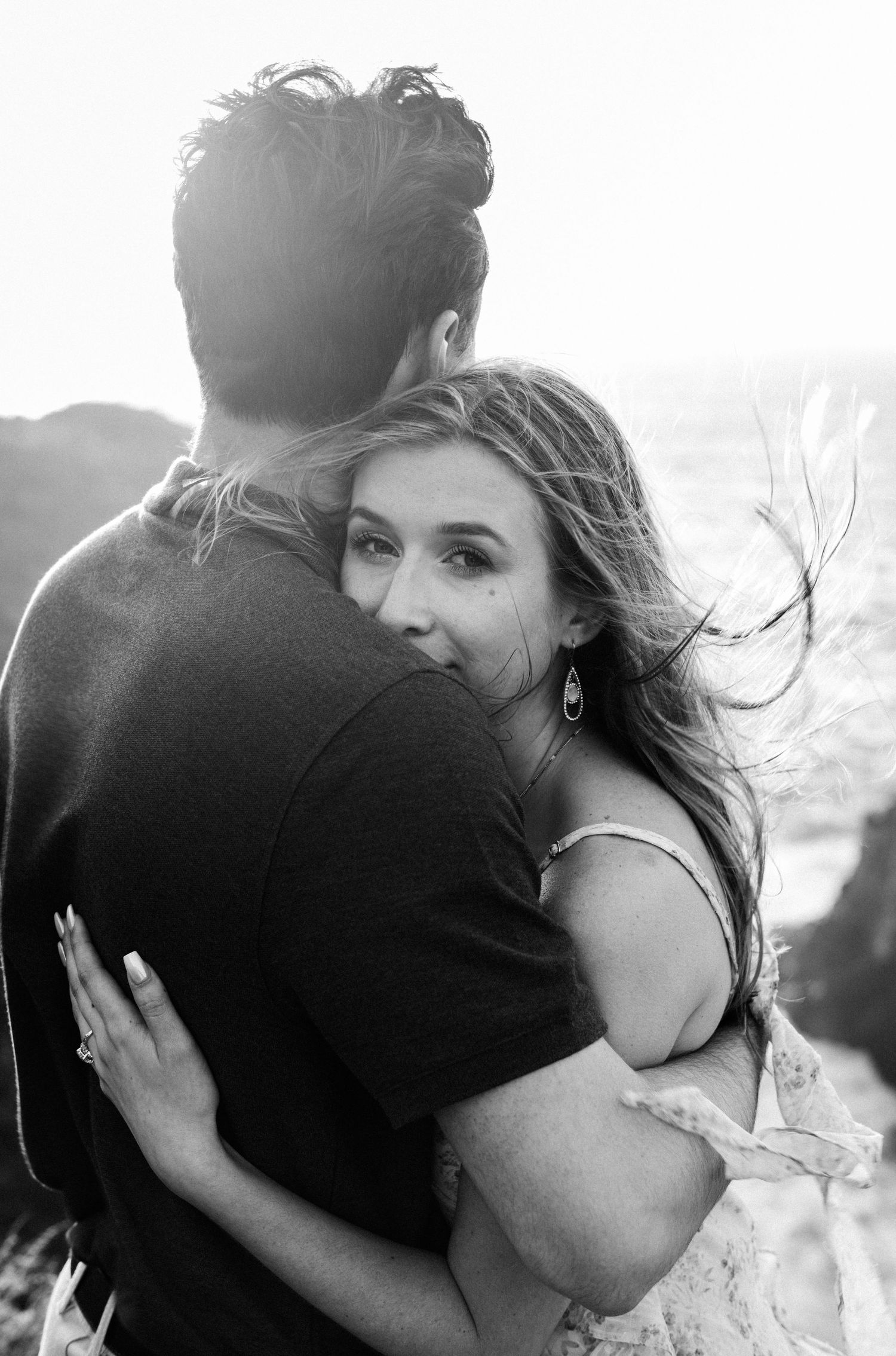 Black and white photo of a couple embracing outdoors, woman looking back at camera with windswept hair in golden backlight.