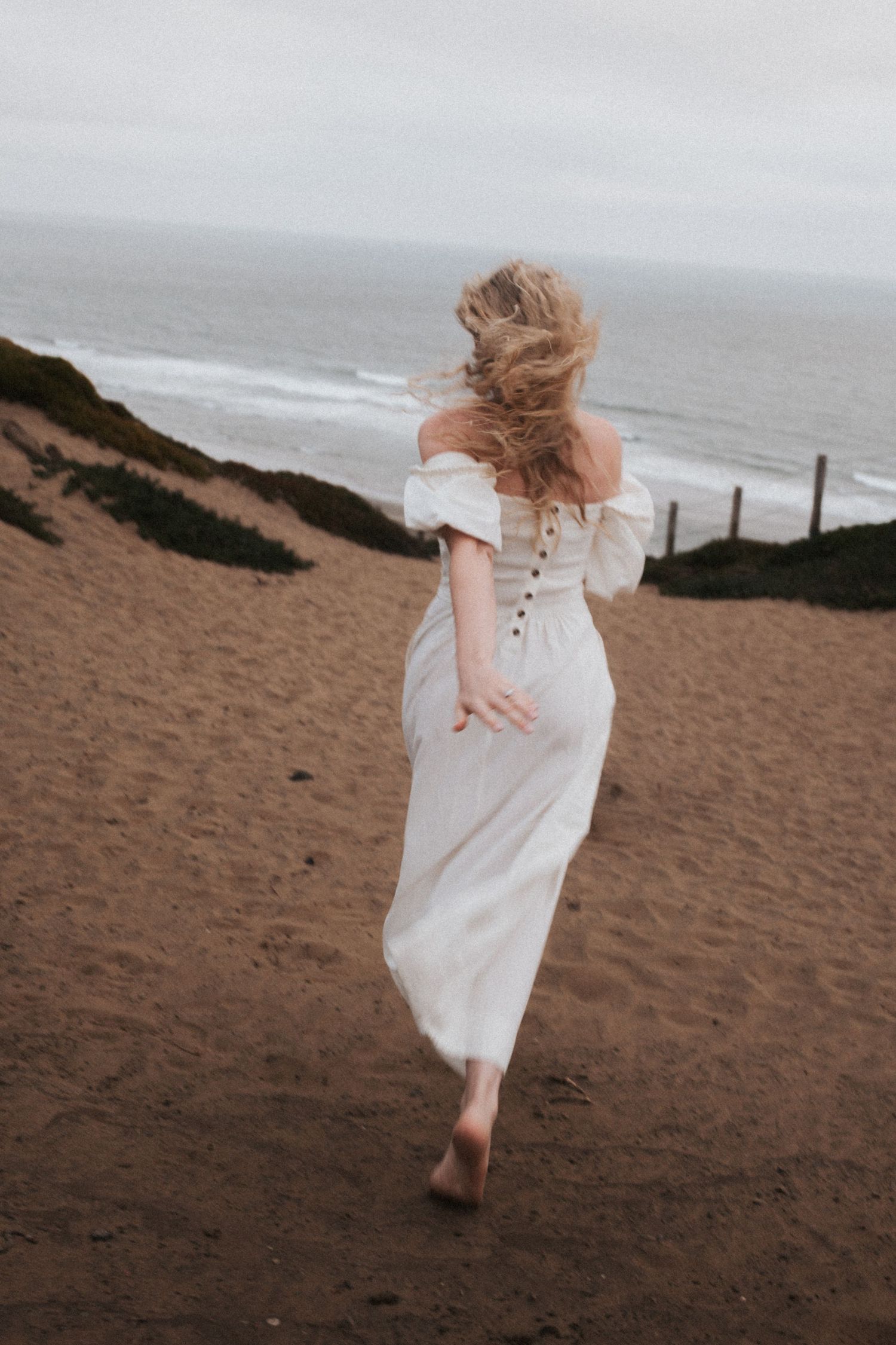 A barefoot woman in a flowing white off-shoulder dress runs along a sandy coastal path on a cloudy day.