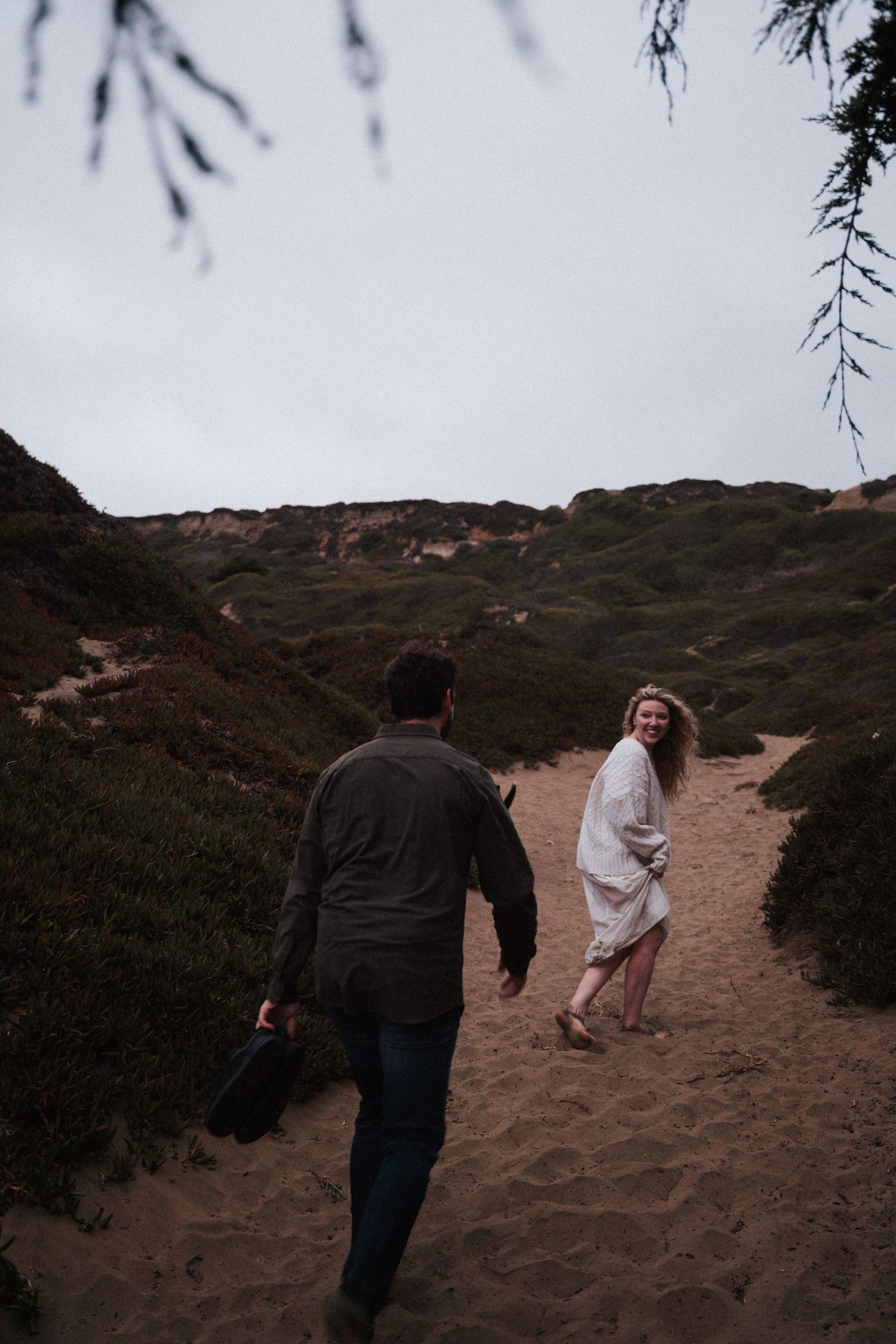 A smiling woman in white looks back at her partner while hiking up a sandy coastal trail.