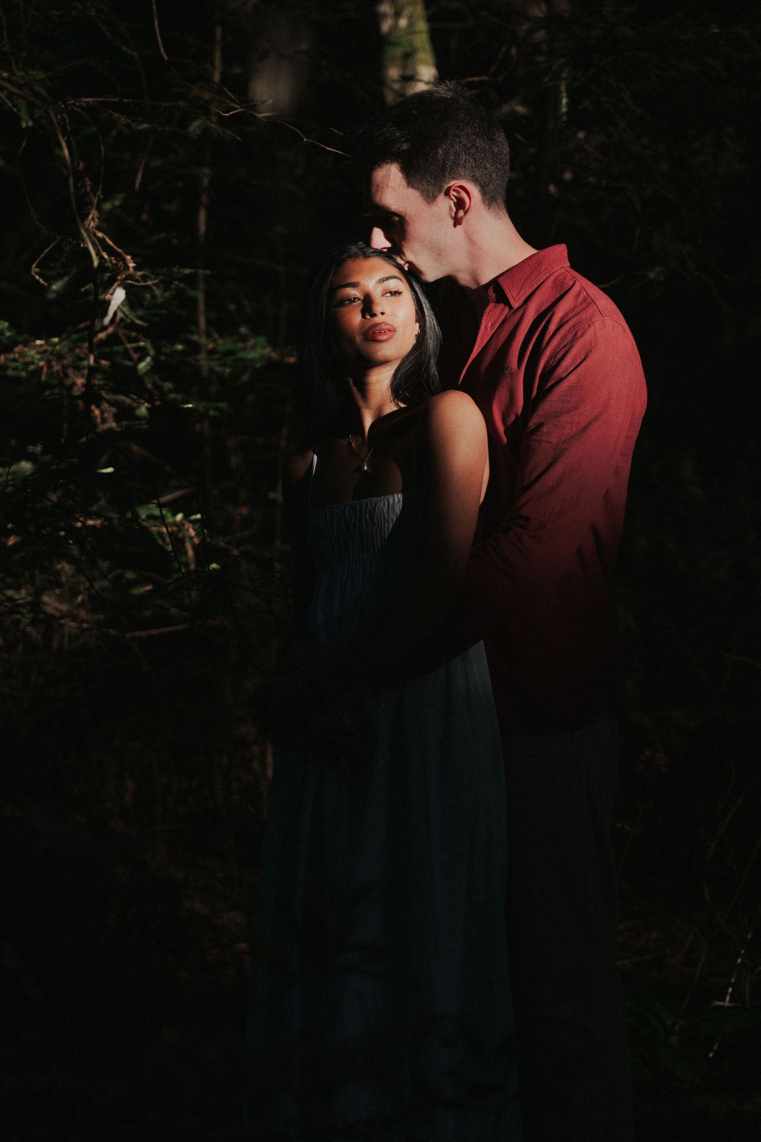 Couple embracing in a dark forest, woman in teal dress and man in red shirt sharing a tender moment.Photography by The Y.K. Studio.