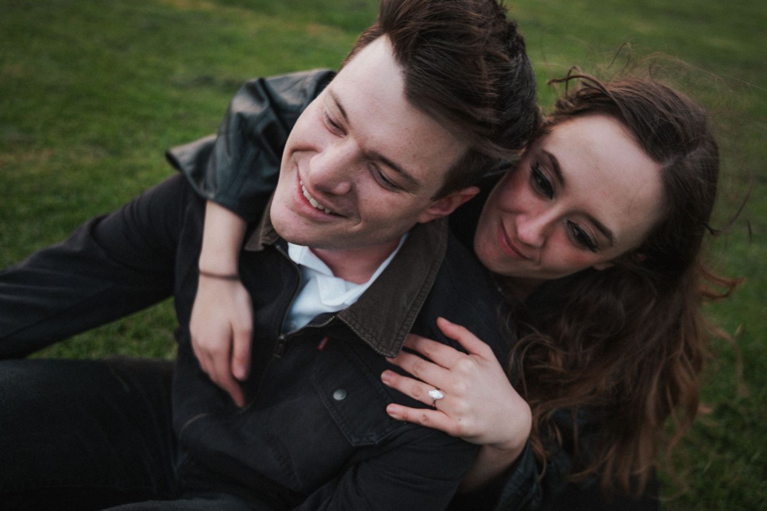Happy couple sitting on grass, woman resting head on smiling man's shoulder in a tender outdoor moment.