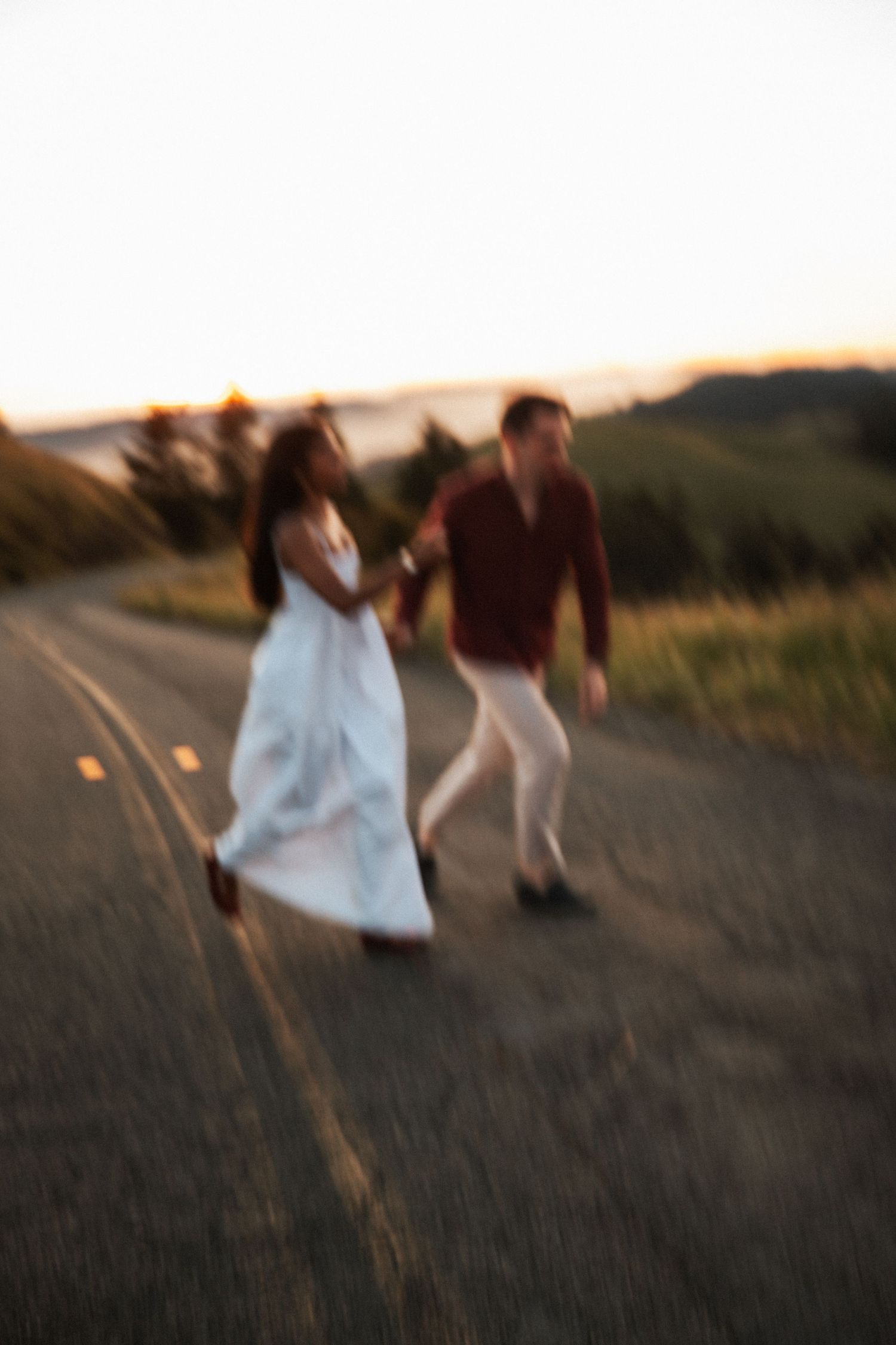Blurry couple holding hands running on a rural road at golden hour sunset with green hills in background.