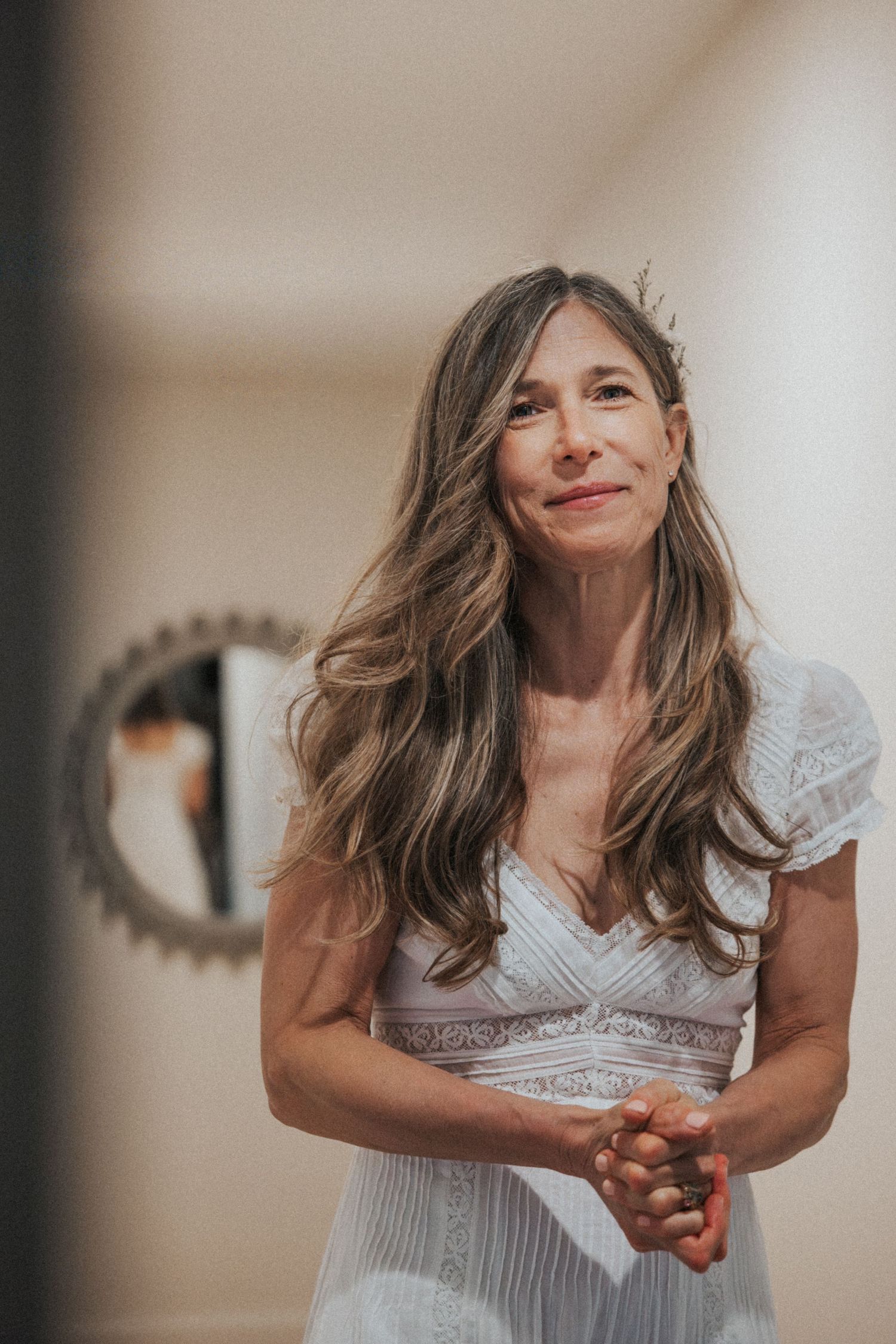 A woman in a white lace dress smiles warmly indoors with long flowing hair.