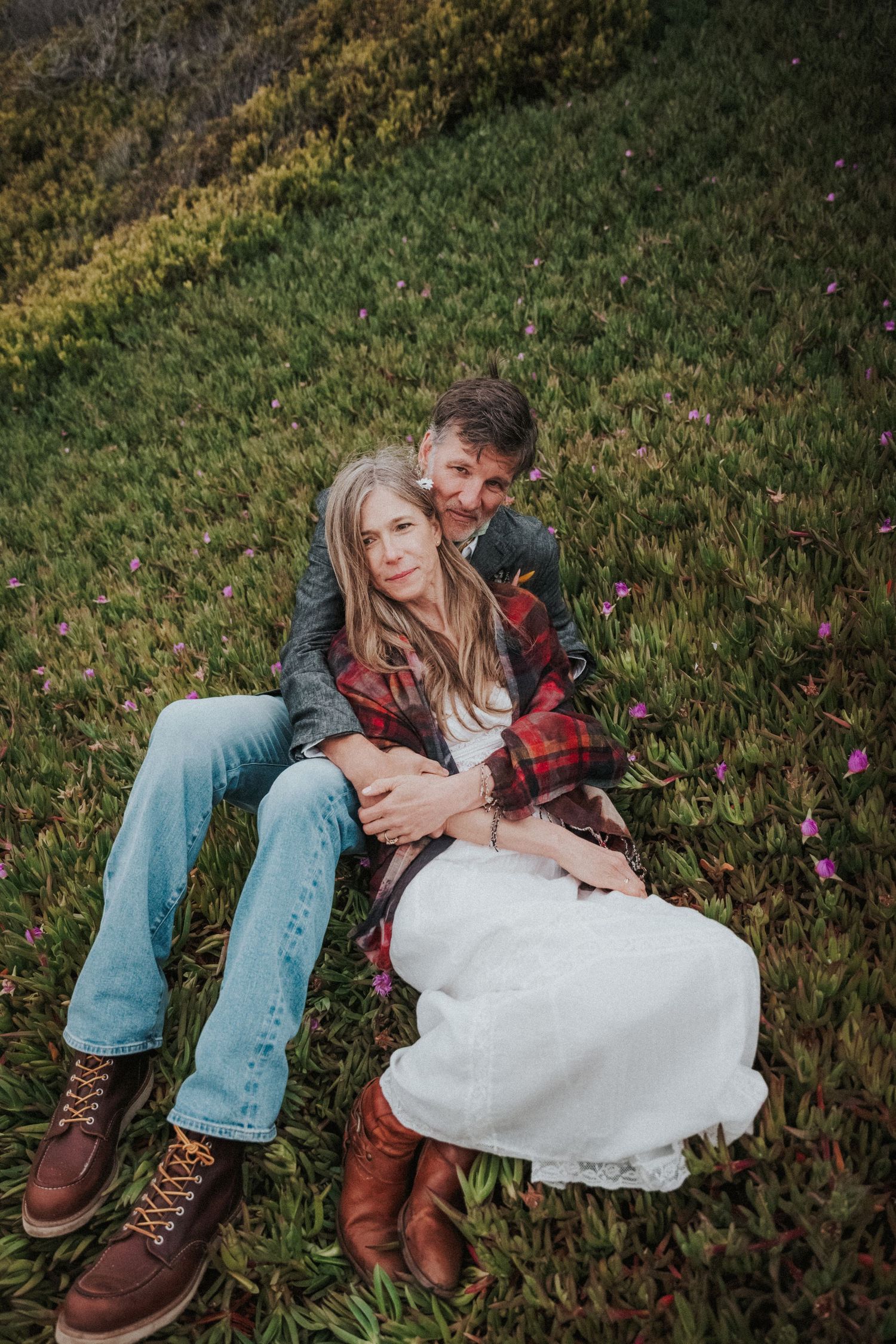 Couple sitting together on a grassy hillside, woman in plaid jacket and jeans embracing tenderly.