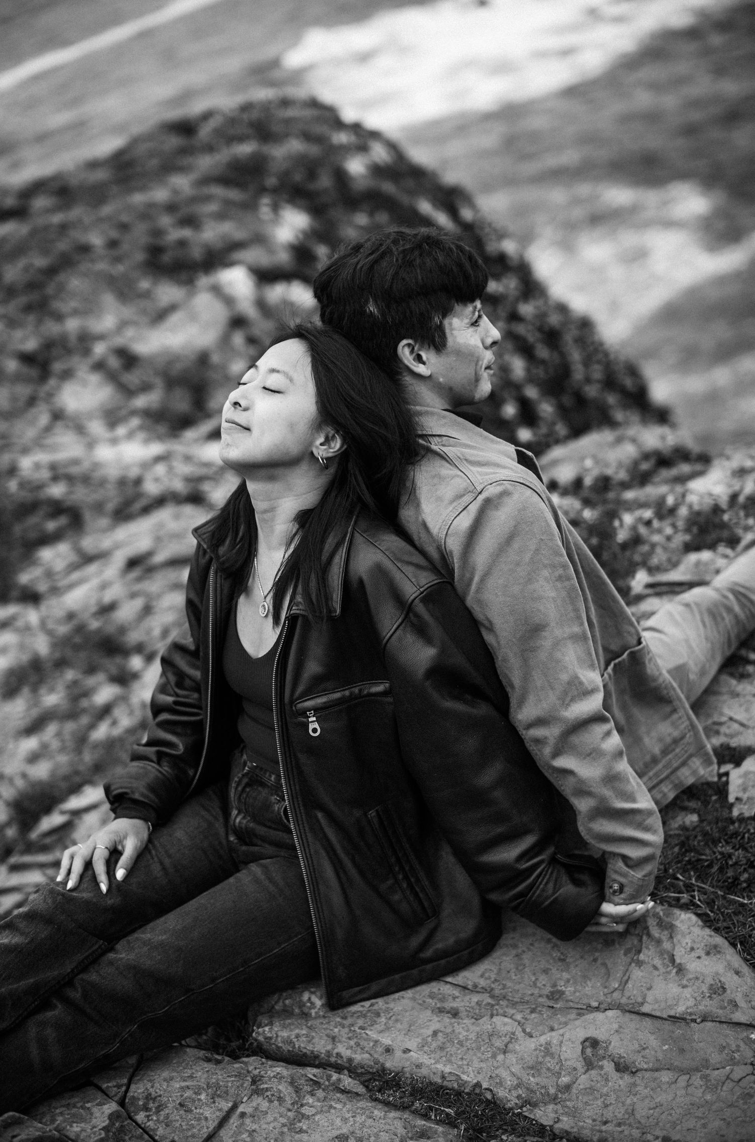 Black and white photo of couple sitting back to back on rocks outdoors.