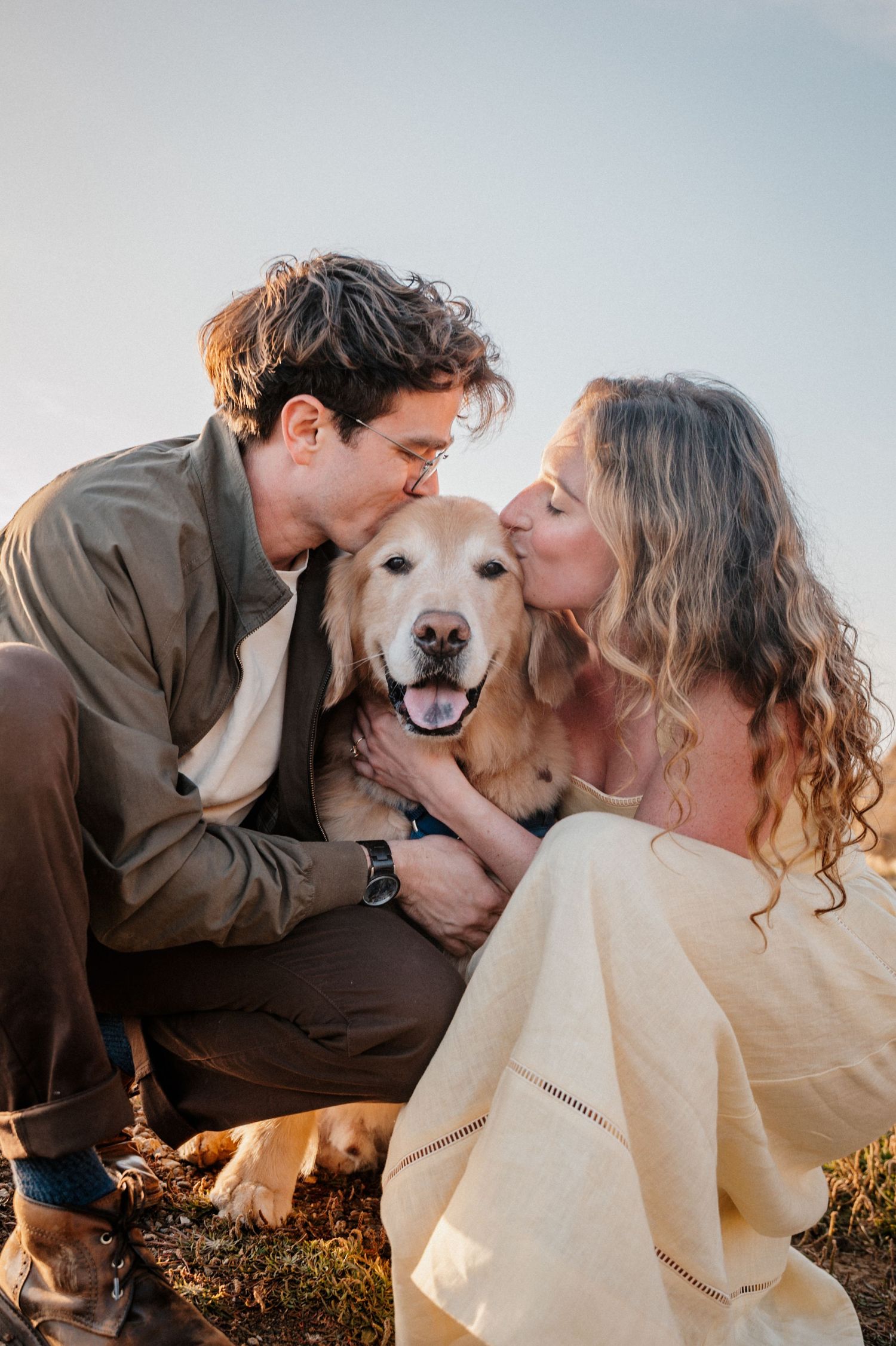 A happy couple kisses their smiling golden retriever dog outdoors at sunset.