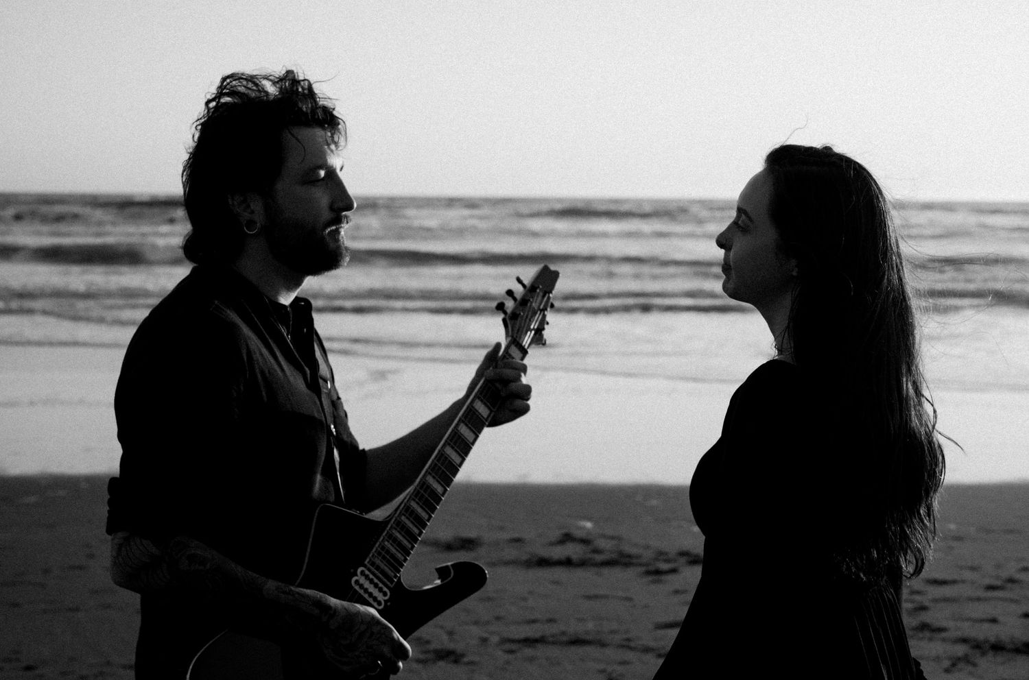 Man playing electric guitar serenading woman on beach at dusk in black and white.