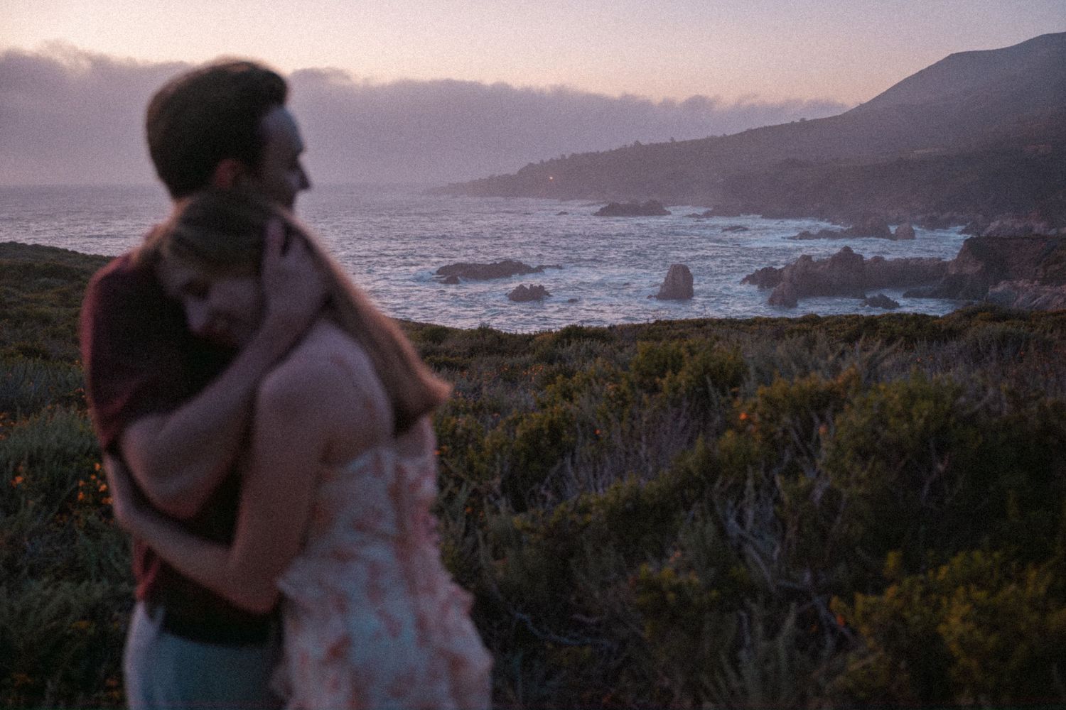 Couple embracing at sunset on a rocky coastal cliff with misty ocean views and dramatic twilight sky.