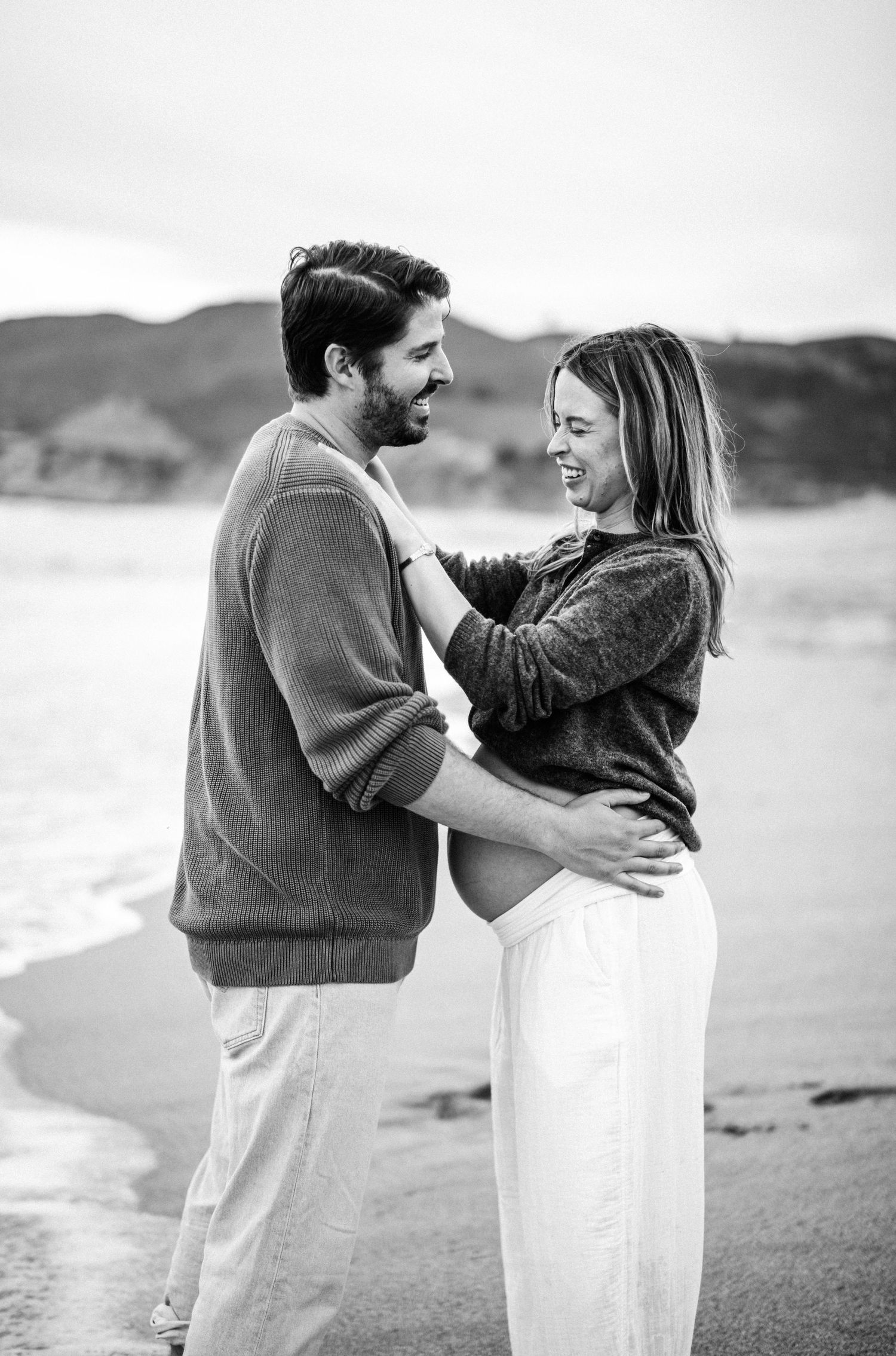 Black and white beach maternity photo of a smiling couple embracing, with ocean waves and hills in the background.
