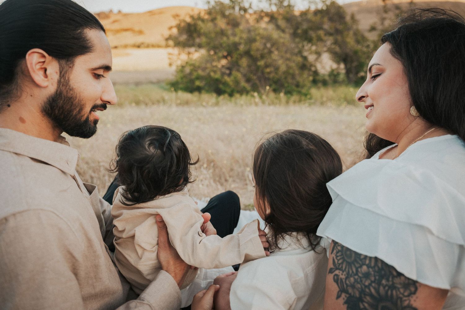 A loving family of four sits together in a golden field, parents smiling at their two young children.