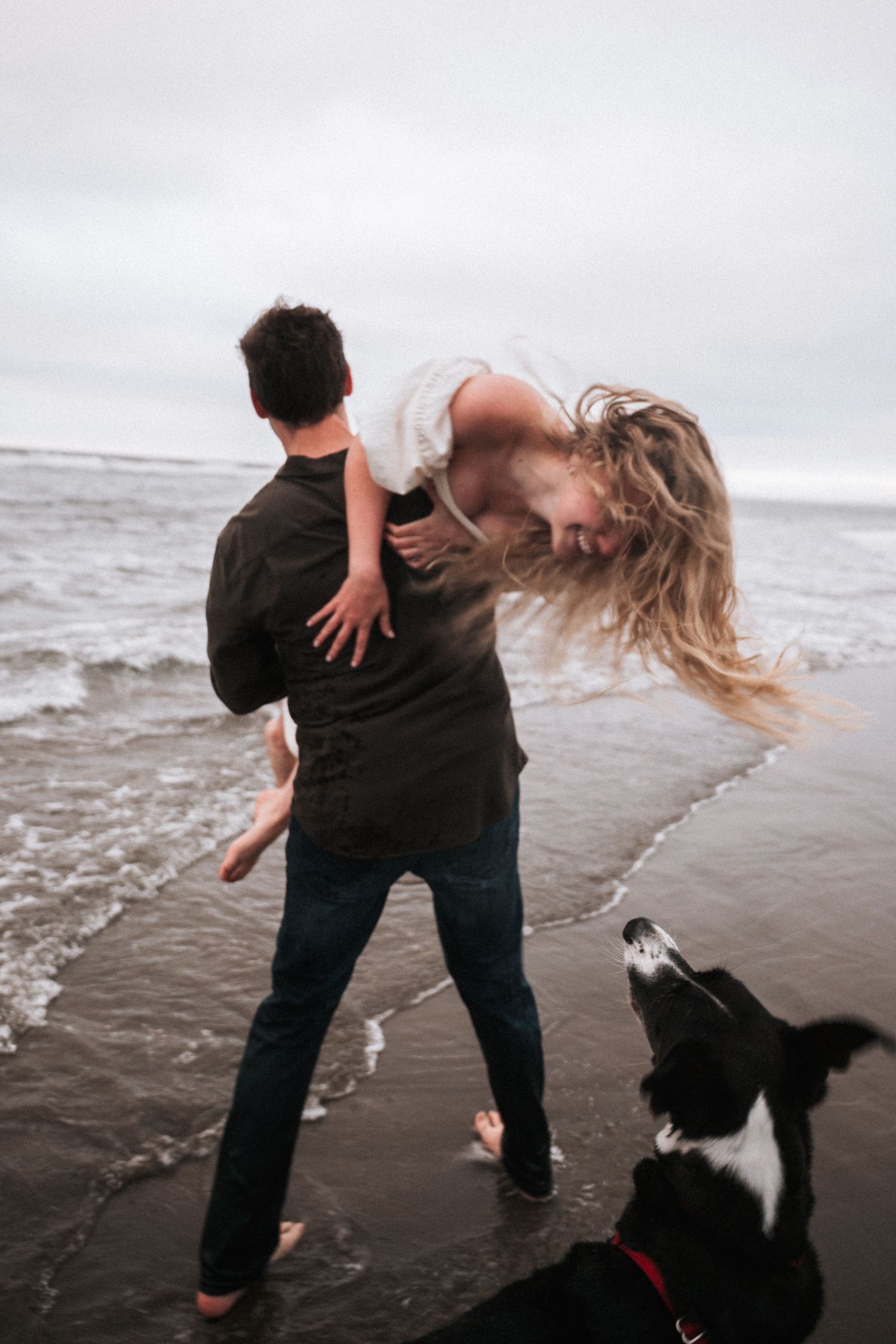 Couple playfully spinning on a dark sandy beach with a black and white dog nearby on an overcast day.