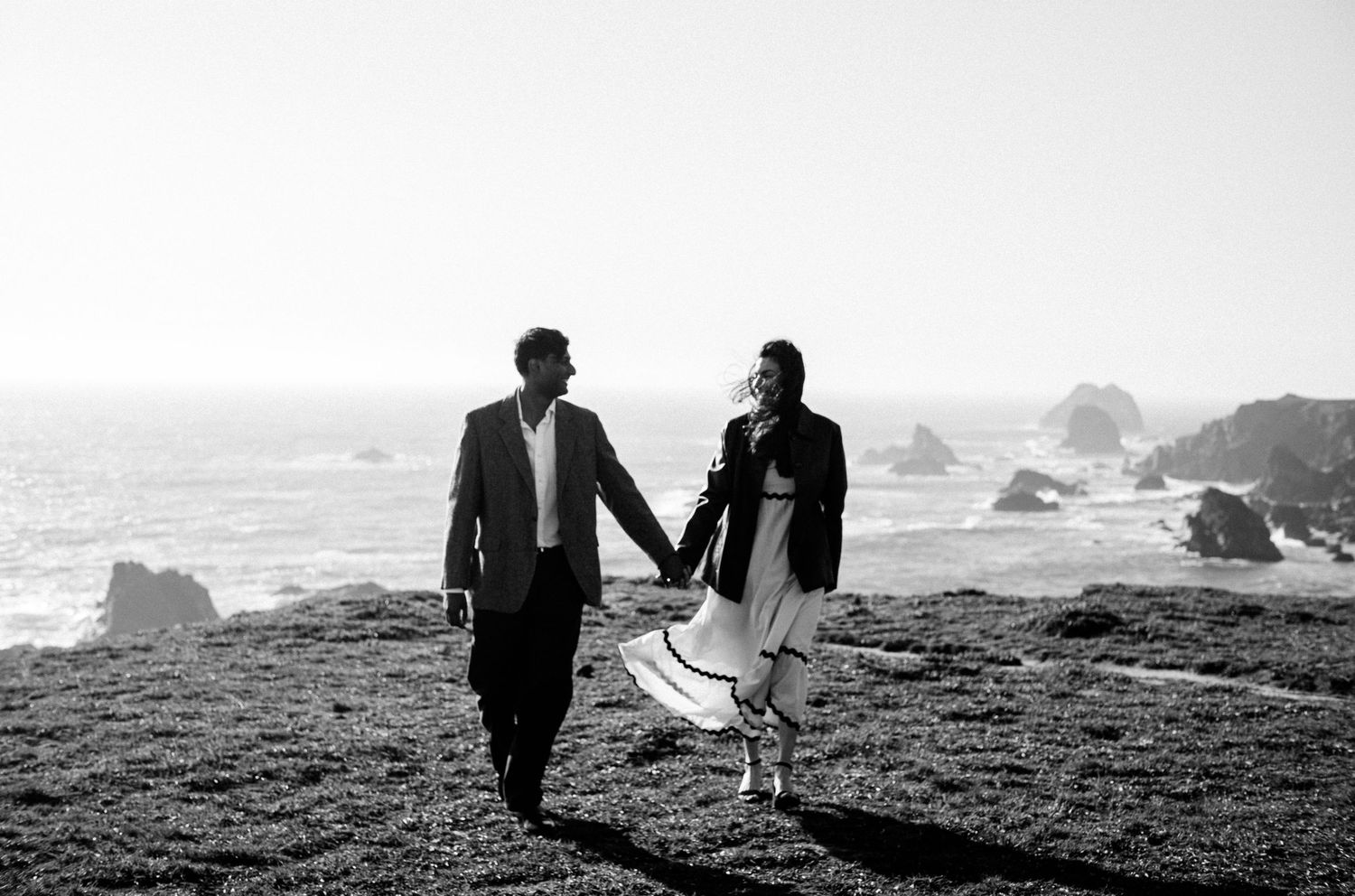 Black and white photo of a couple holding hands walking along a scenic coastal cliffside on their wedding day.