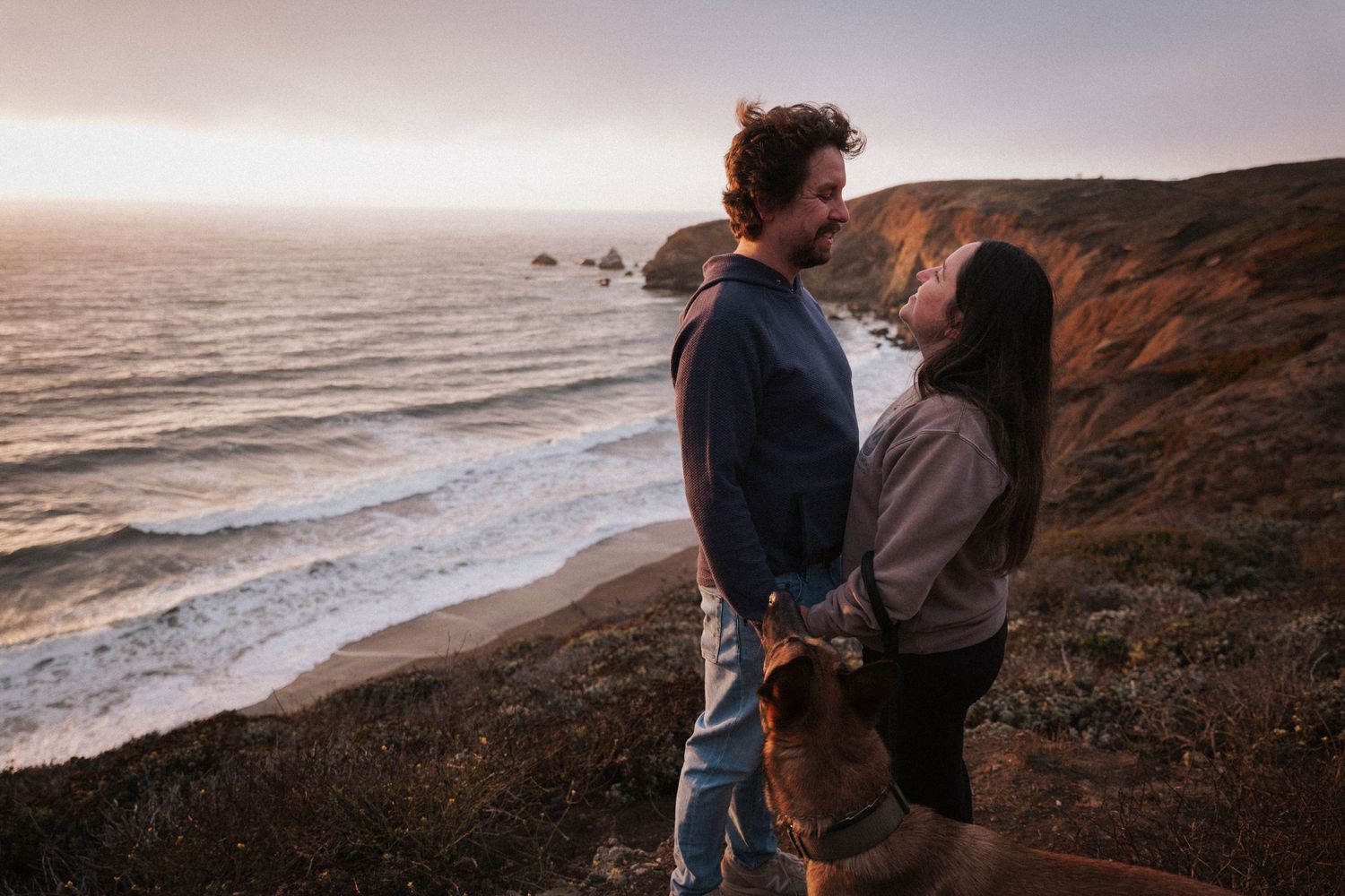 Couple embracing on coastal cliffside at sunset with German Shepherd dog and ocean waves in background.