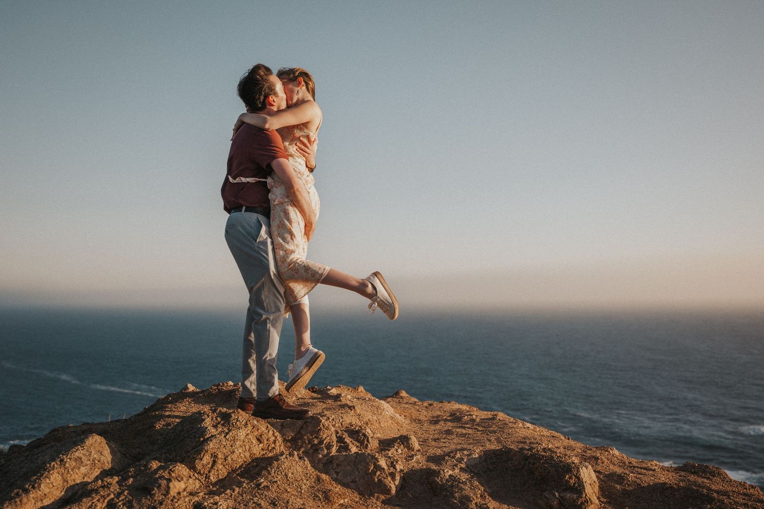 Couple embracing on rocky cliff overlooking ocean at sunset, romantic outdoor engagement photo.