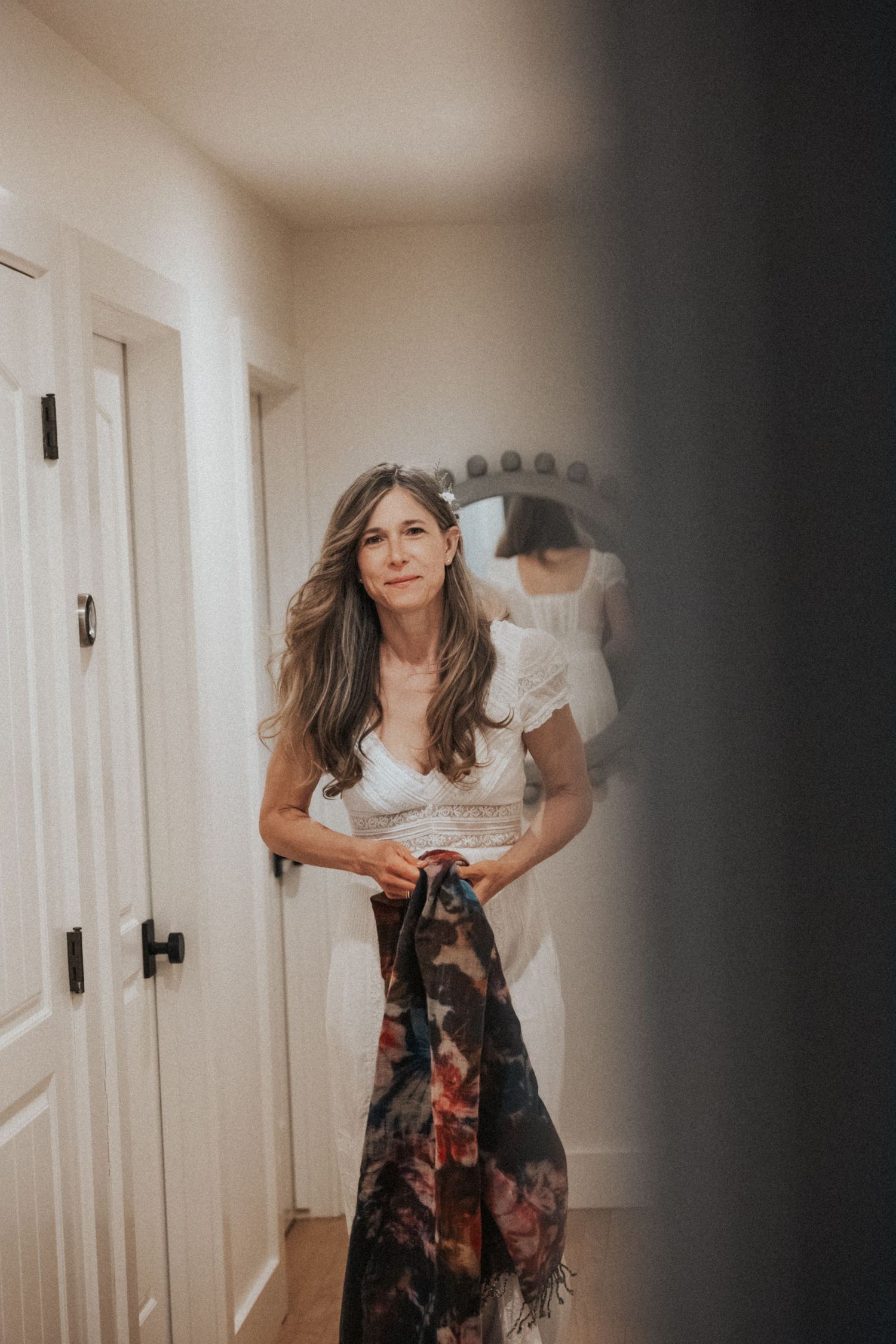 Elegant woman in white lace top holds a floral scarf while standing in a hallway with a mirror reflection behind her.