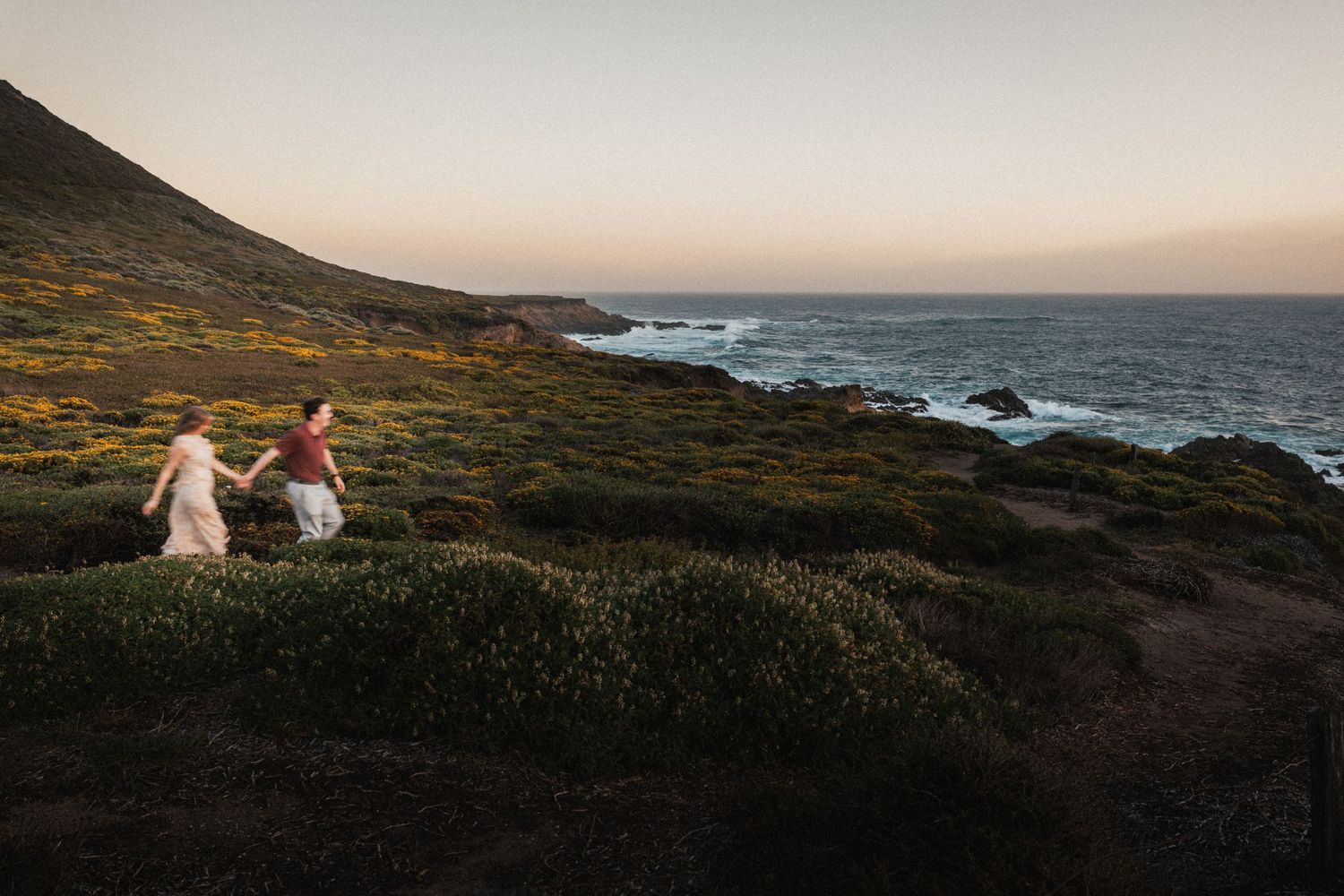 Couple holding hands running along a scenic coastal hillside at sunset with ocean waves in the background.