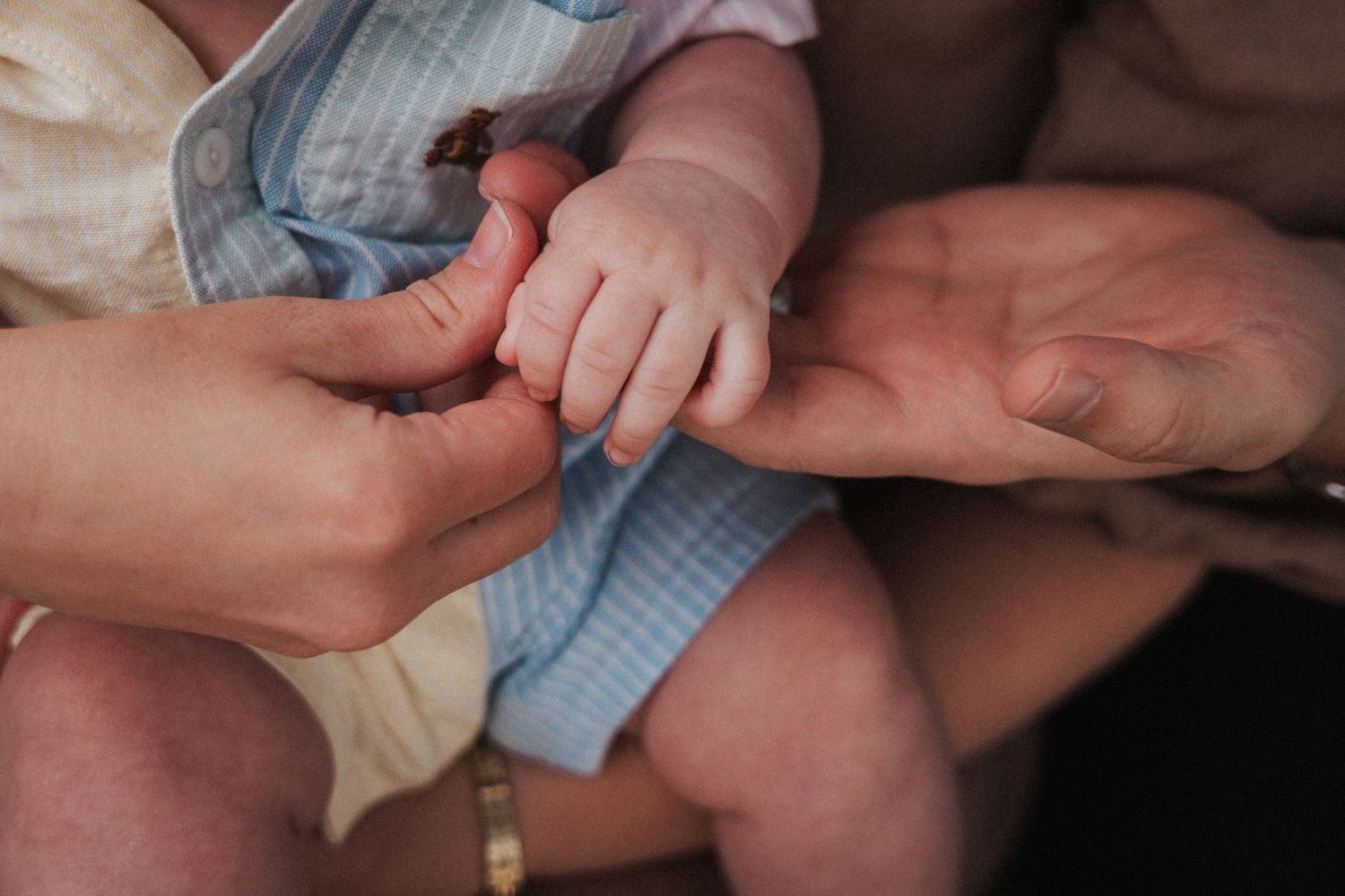 Close-up of a newborn baby's tiny hand grasping adult fingers, wearing a light blue outfit.