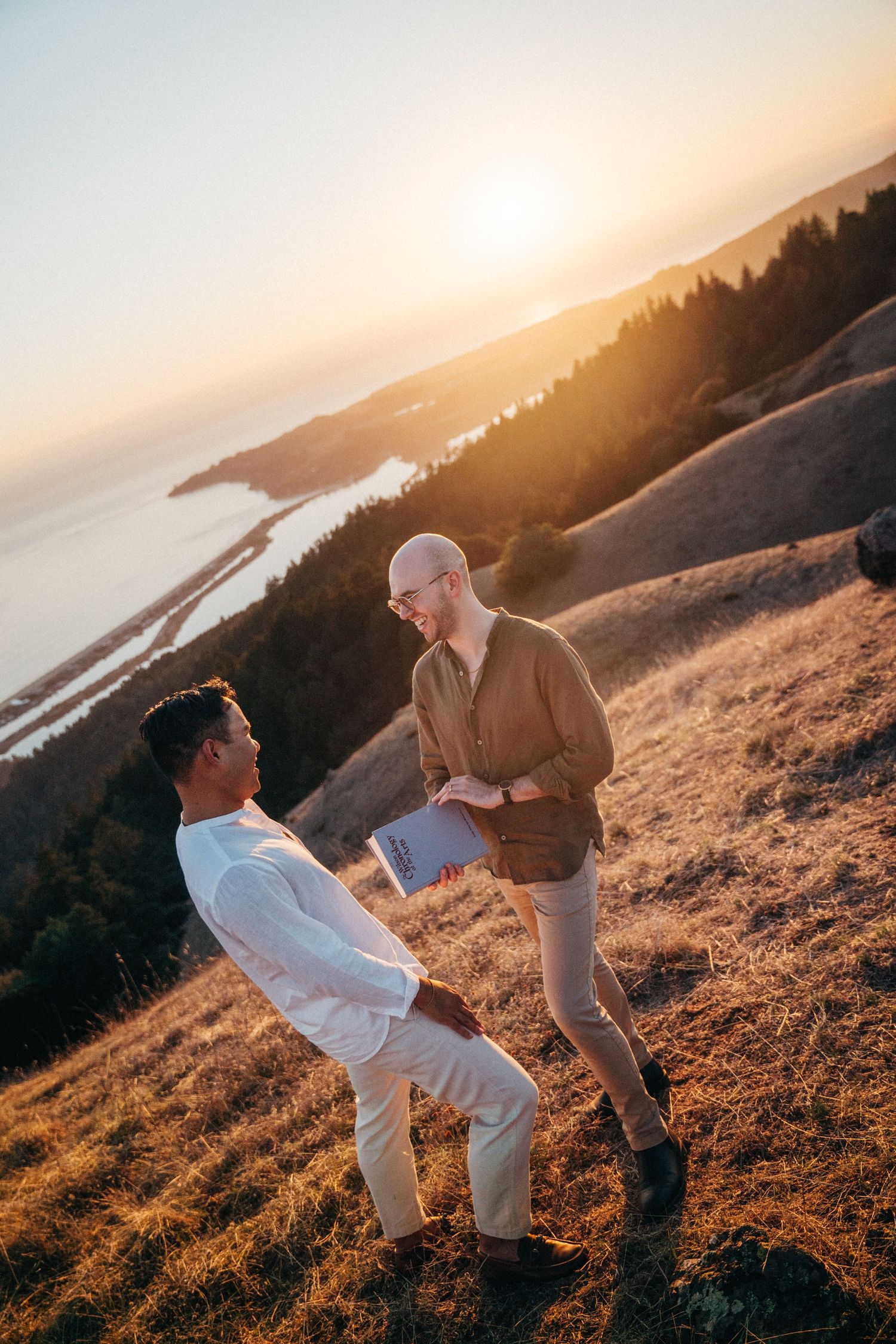 Two men share a romantic moment on a sunlit hillside with a stunning coastal sunset view behind them.