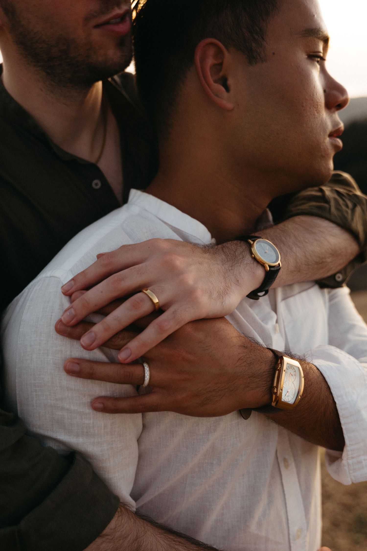 Close-up of two men embracing, wearing rings and watches at sunset.